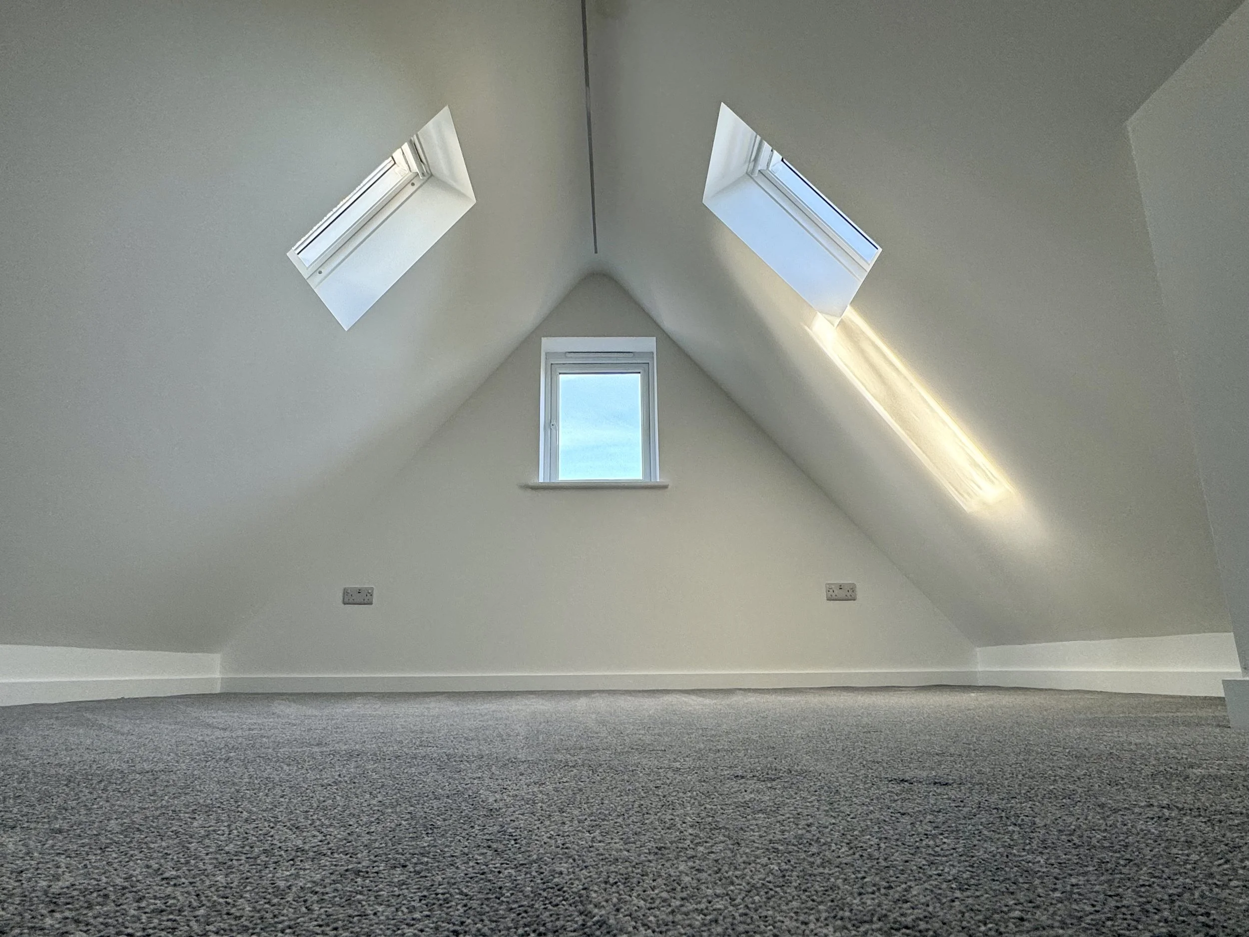 Empty attic room with sloped ceiling, three skylight windows, white walls, gray carpeted floor, and electrical outlets.