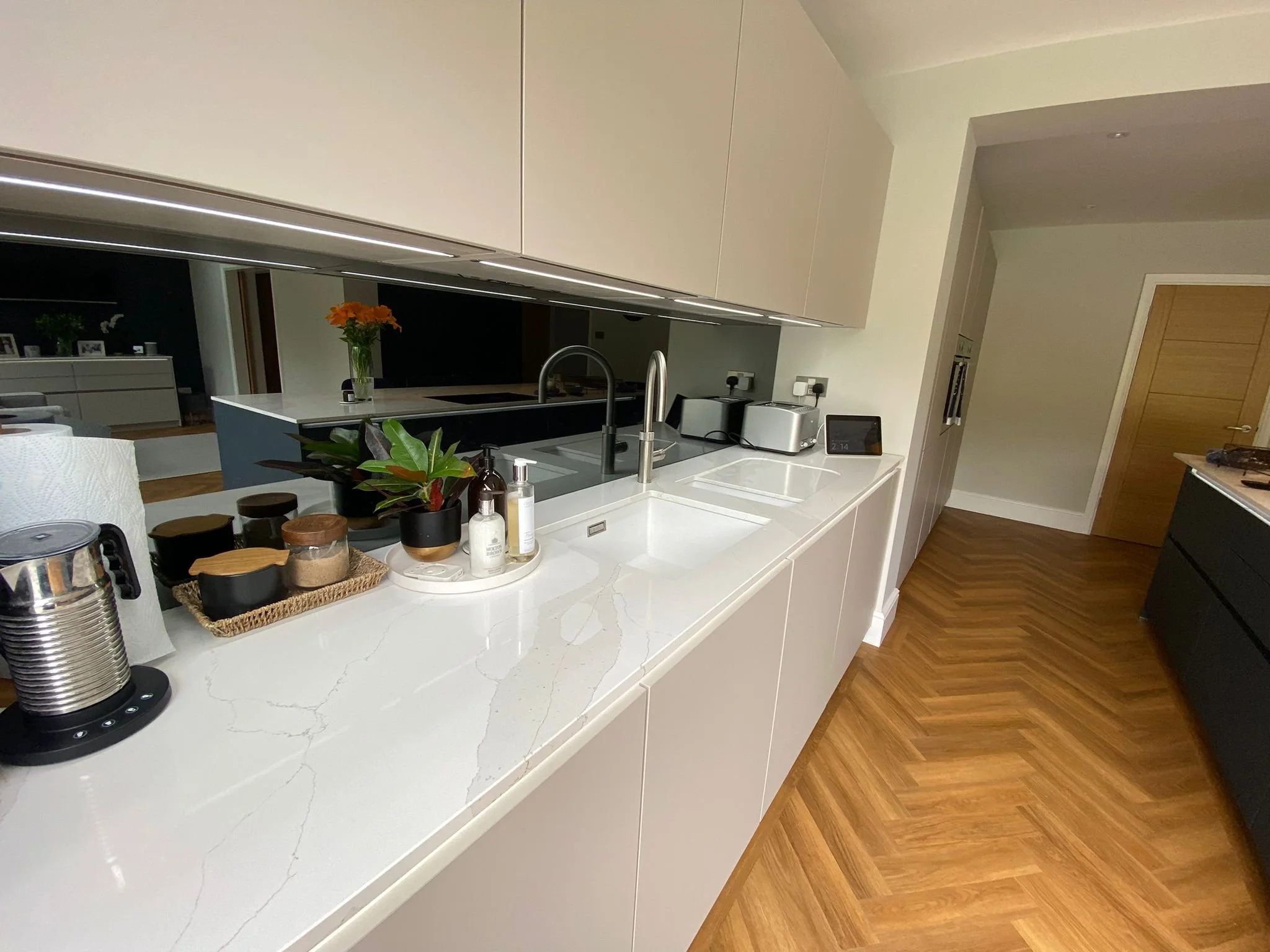 Modern kitchen with white countertops, built-in sink, plants, jars, and kitchen appliances, with wooden flooring and beige cabinets.