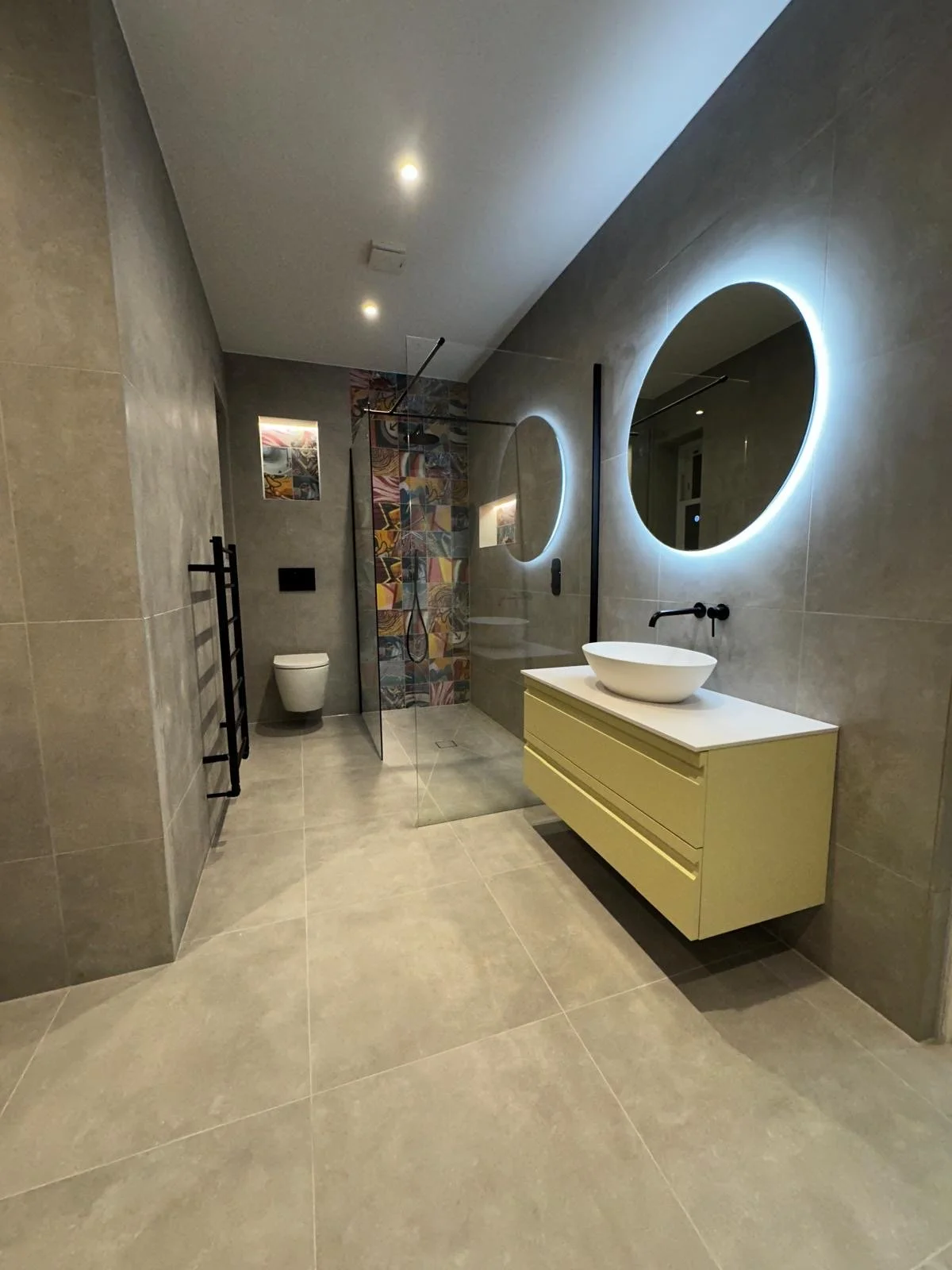 Modern bathroom with beige tiles, circular backlit mirrors, floating vanity with vessel sink, walk-in shower with glass partition, and wall-mounted toilet.