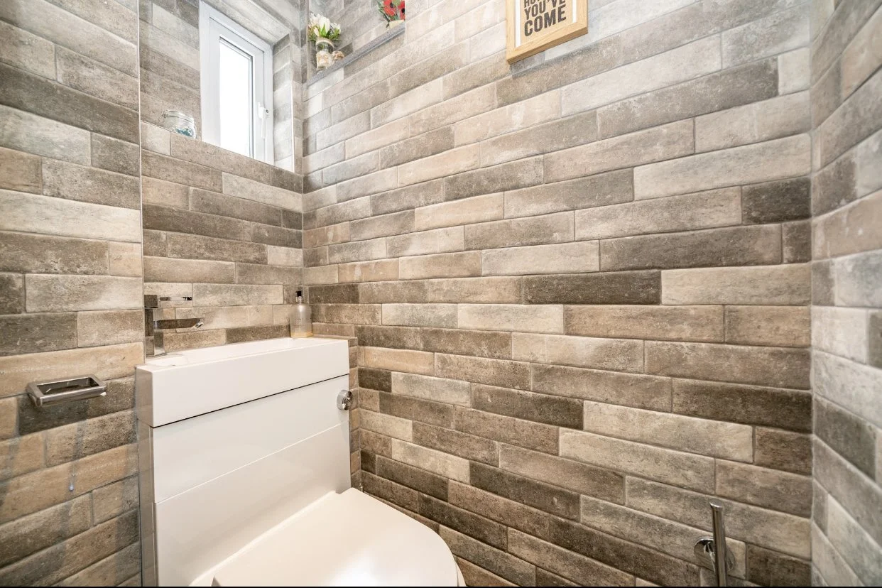 Small bathroom with beige brick wall tiles, a white tankless toilet, a small window with potted plants, and a framed sign on the wall.