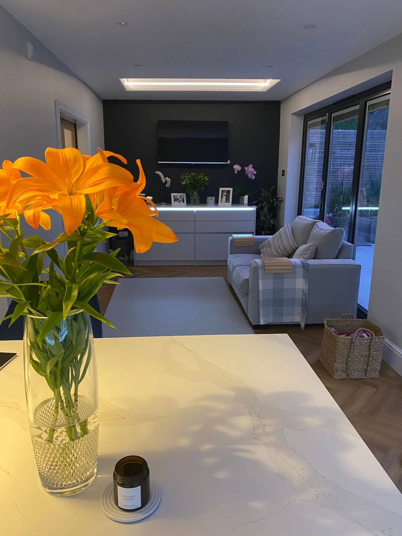 Living room with beige couch, white cabinet, wall-mounted TV, sliding glass door, and a vase with orange lilies on a white countertop.