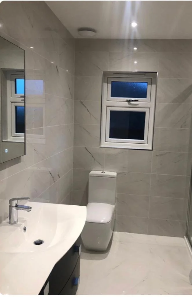 Modern bathroom with beige marble tiles, a white toilet, a white curved sink with a silver faucet, a wall-mounted mirror, and a frosted glass window.