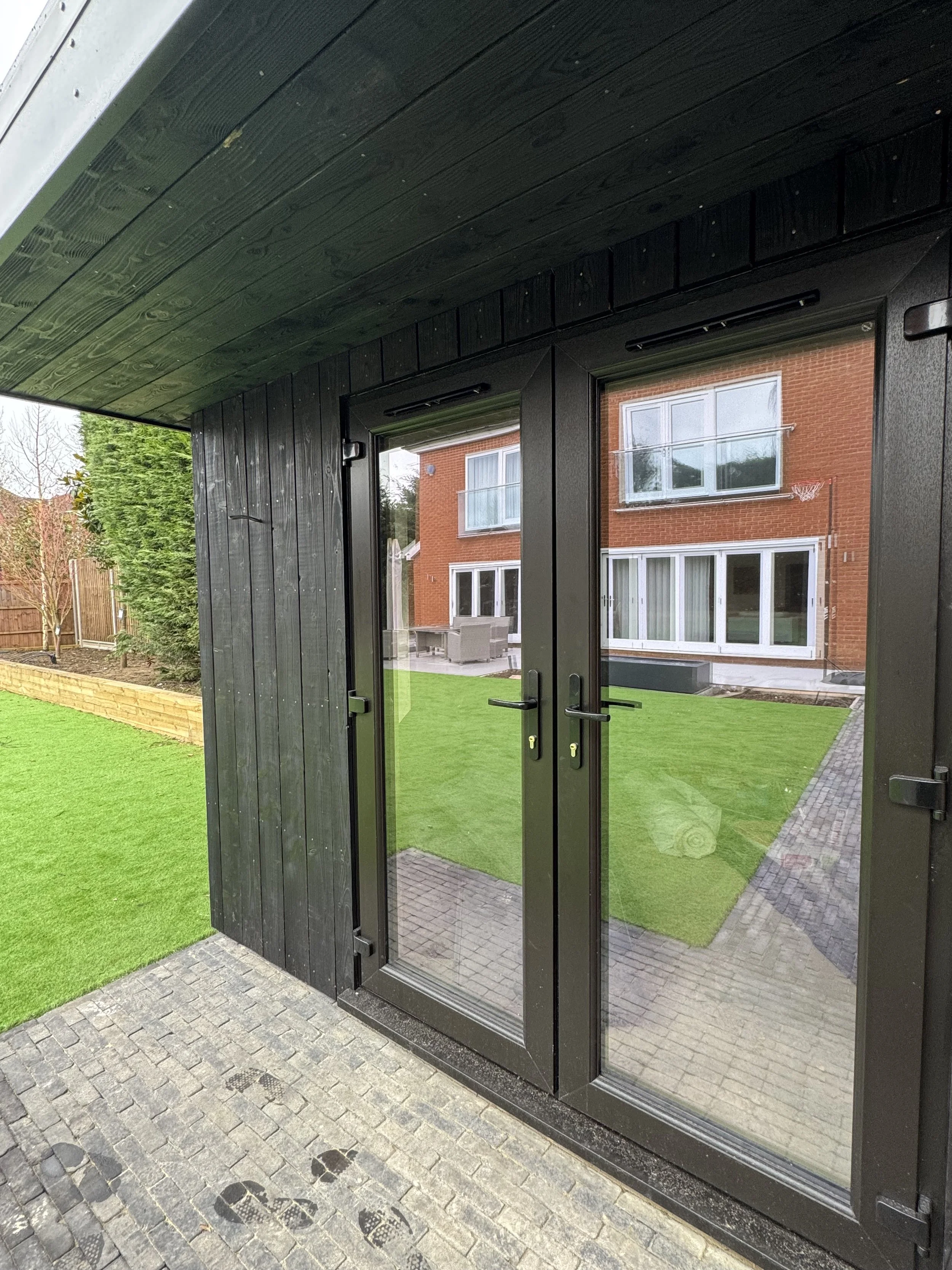 Black-framed glass door leading to a backyard with green grass, paved walkway, and a brick house with large windows.