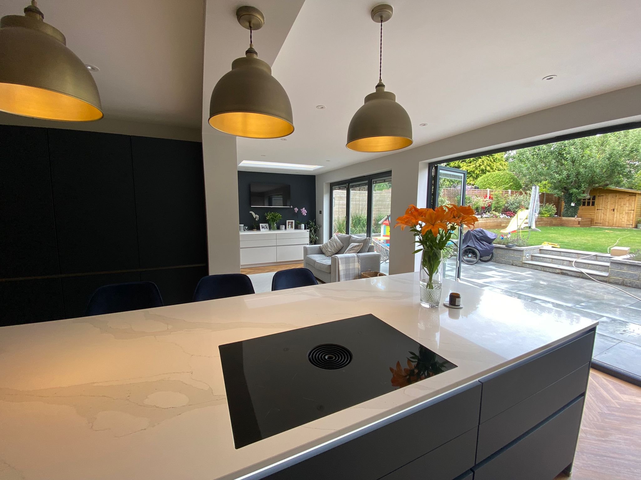 Modern kitchen with white island, black stovetop, three hanging pendant lights, and view of backyard with garden, trees, and shed.
