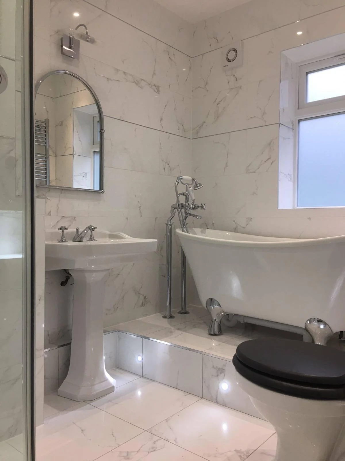Bathroom with marble tiles, pedestal sink, clawfoot bathtub, black toilet seat, and frosted window.