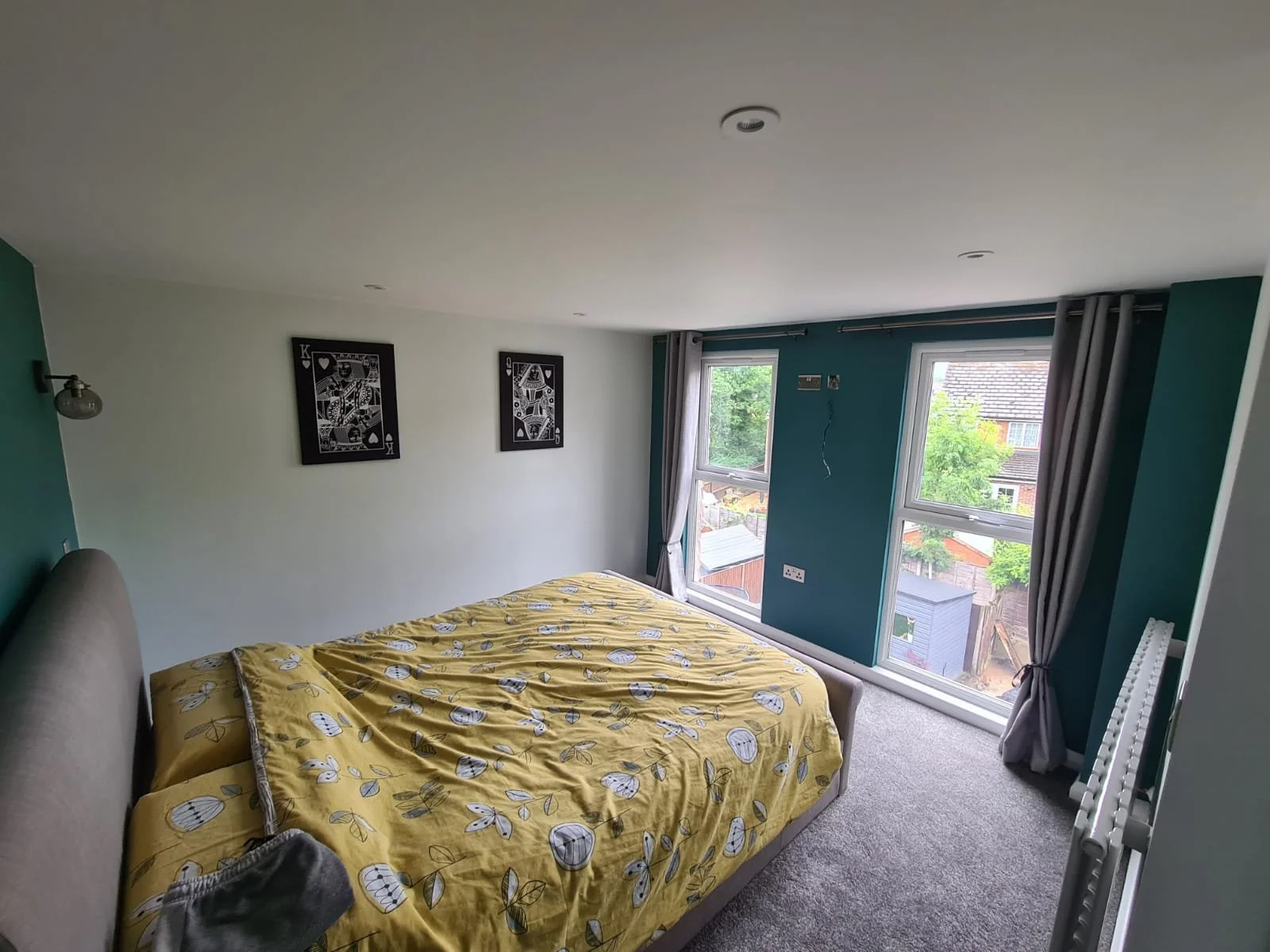 Bedroom with a bed in yellow bedding, dark green and white walls, two black-framed art prints on the white wall, two large windows with gray curtains, and a view of neighboring houses and trees outside.