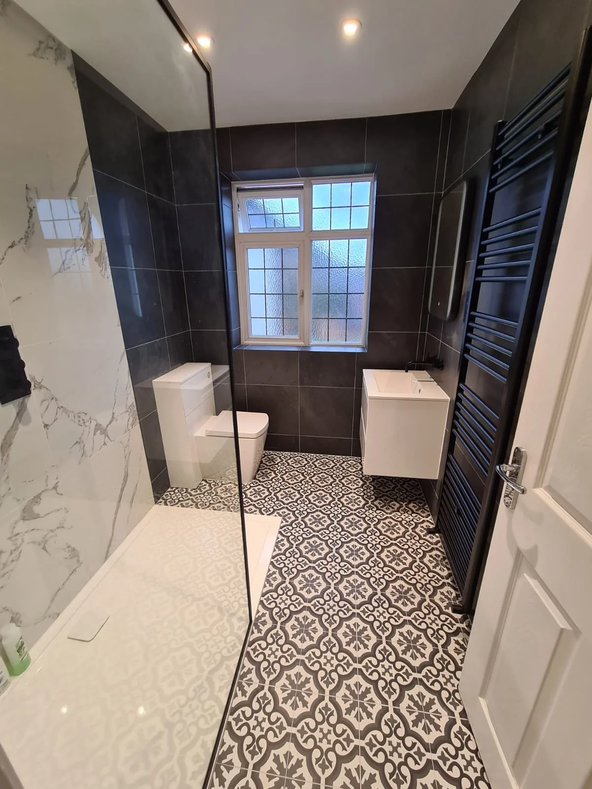 Modern bathroom with black wall tiles, patterned floor, white toilet, small white vanity with sink, frosted window, black towel radiator, and walk-in shower with marble-effect tiles.