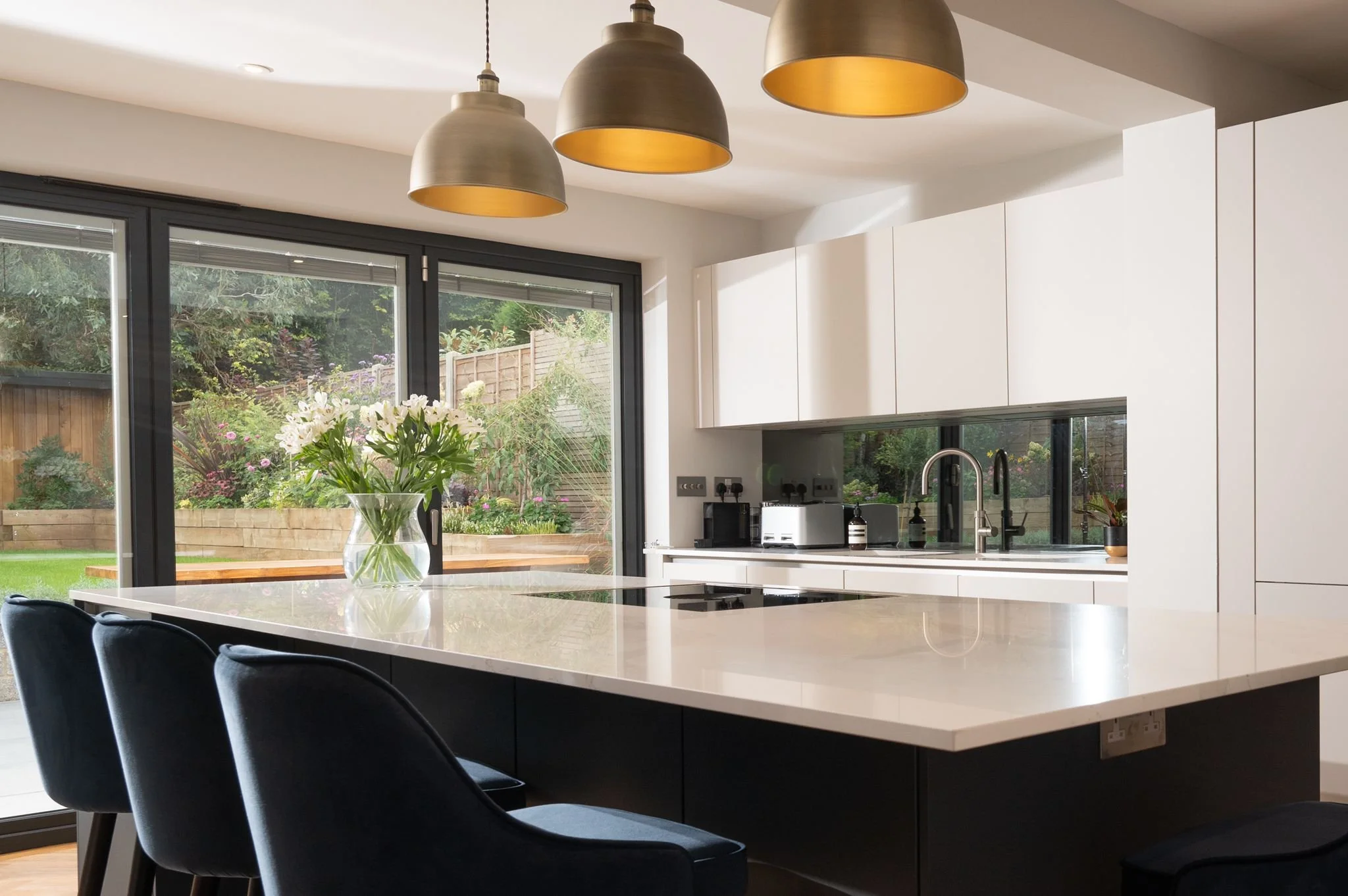 Modern kitchen with white cabinets, large white island, black chairs, pendant lights, and large sliding glass doors opening to a garden with flowers and a wooden fence.