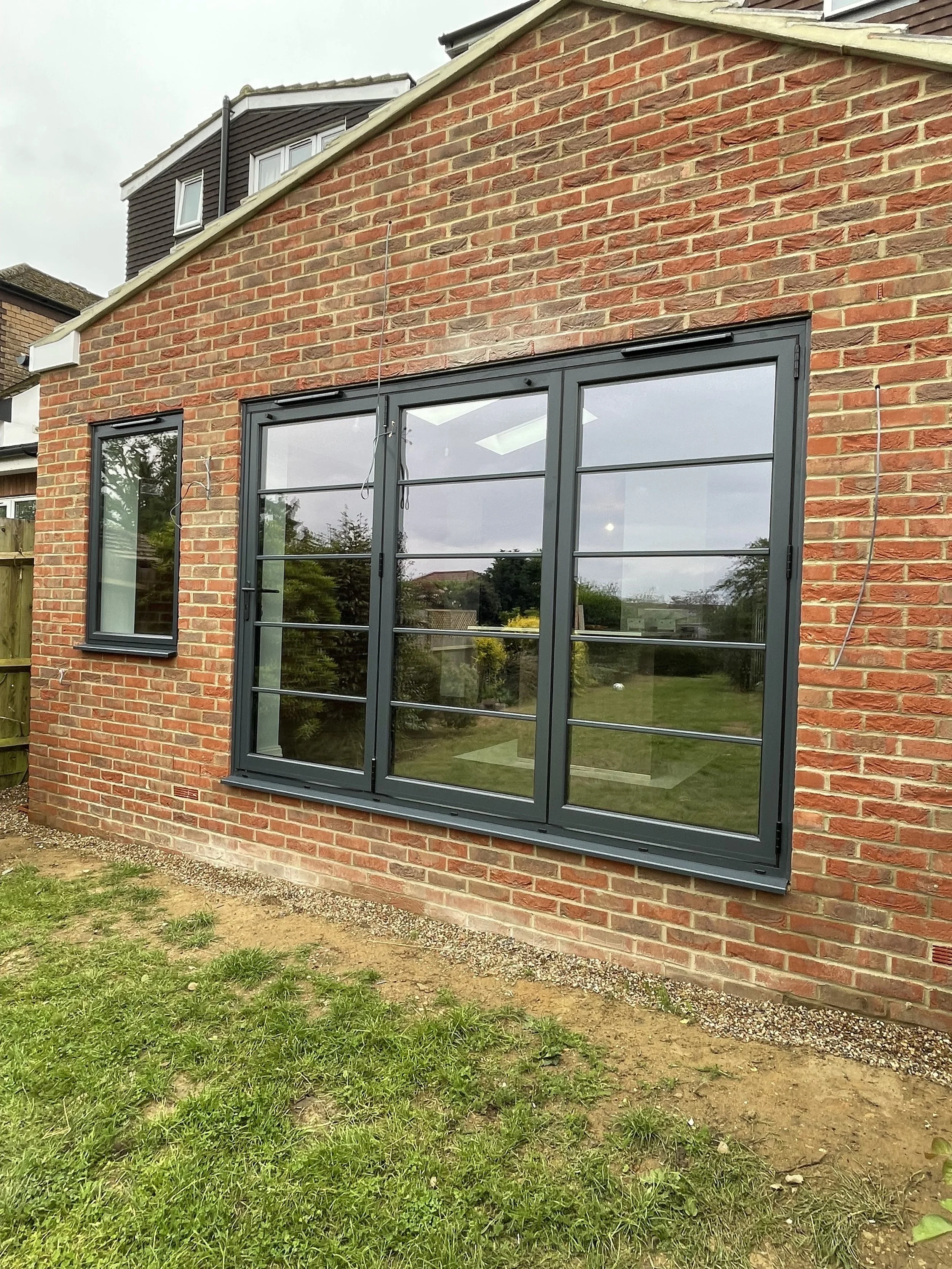 The image shows the exterior of a brick house with a large, modern gray-framed glass window on the wall.