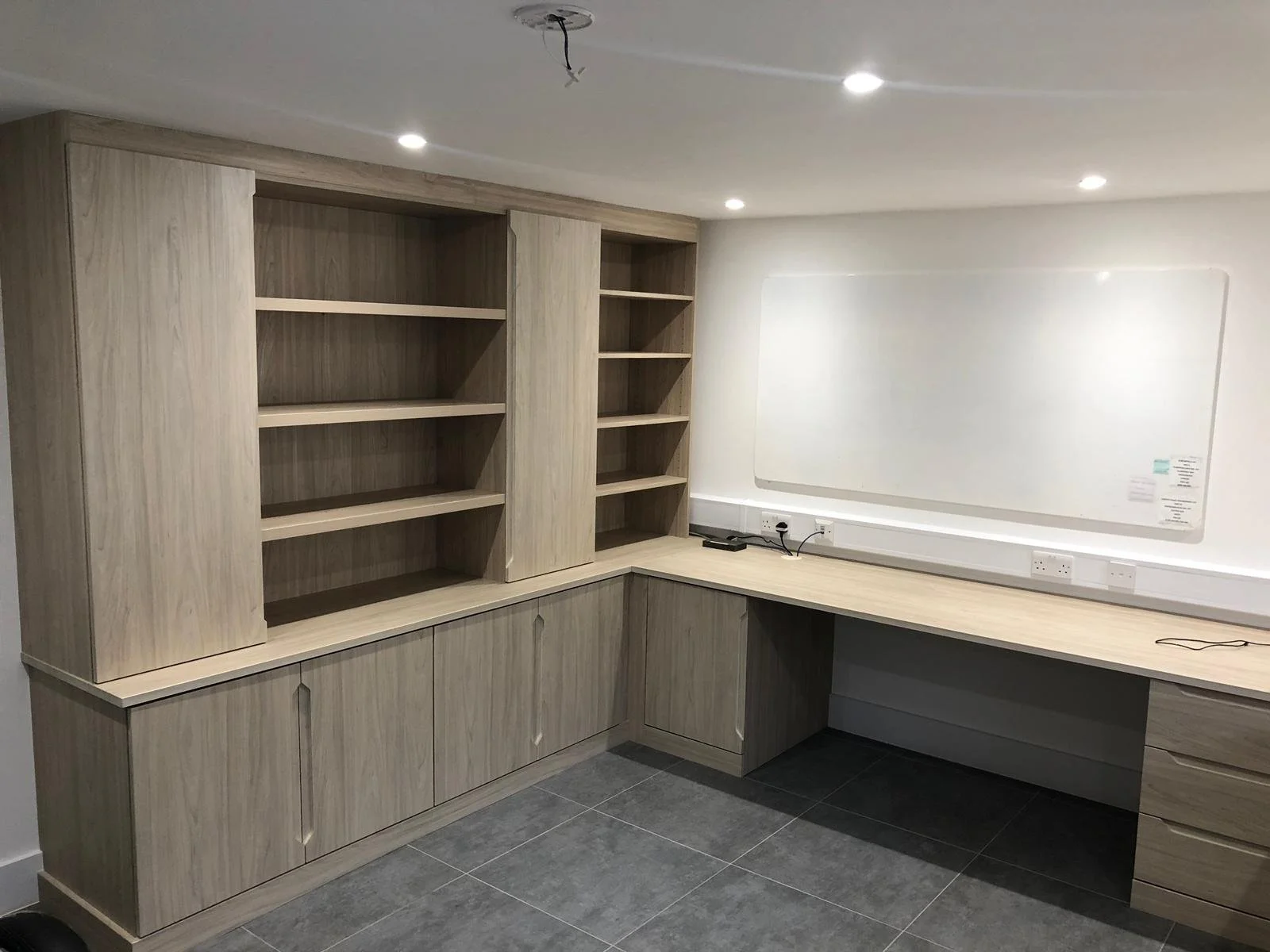 Empty office with light wood built-in shelves and cabinets along the walls, a wooden desk with drawers, a whiteboard on the wall, and a ceiling with recessed lighting.
