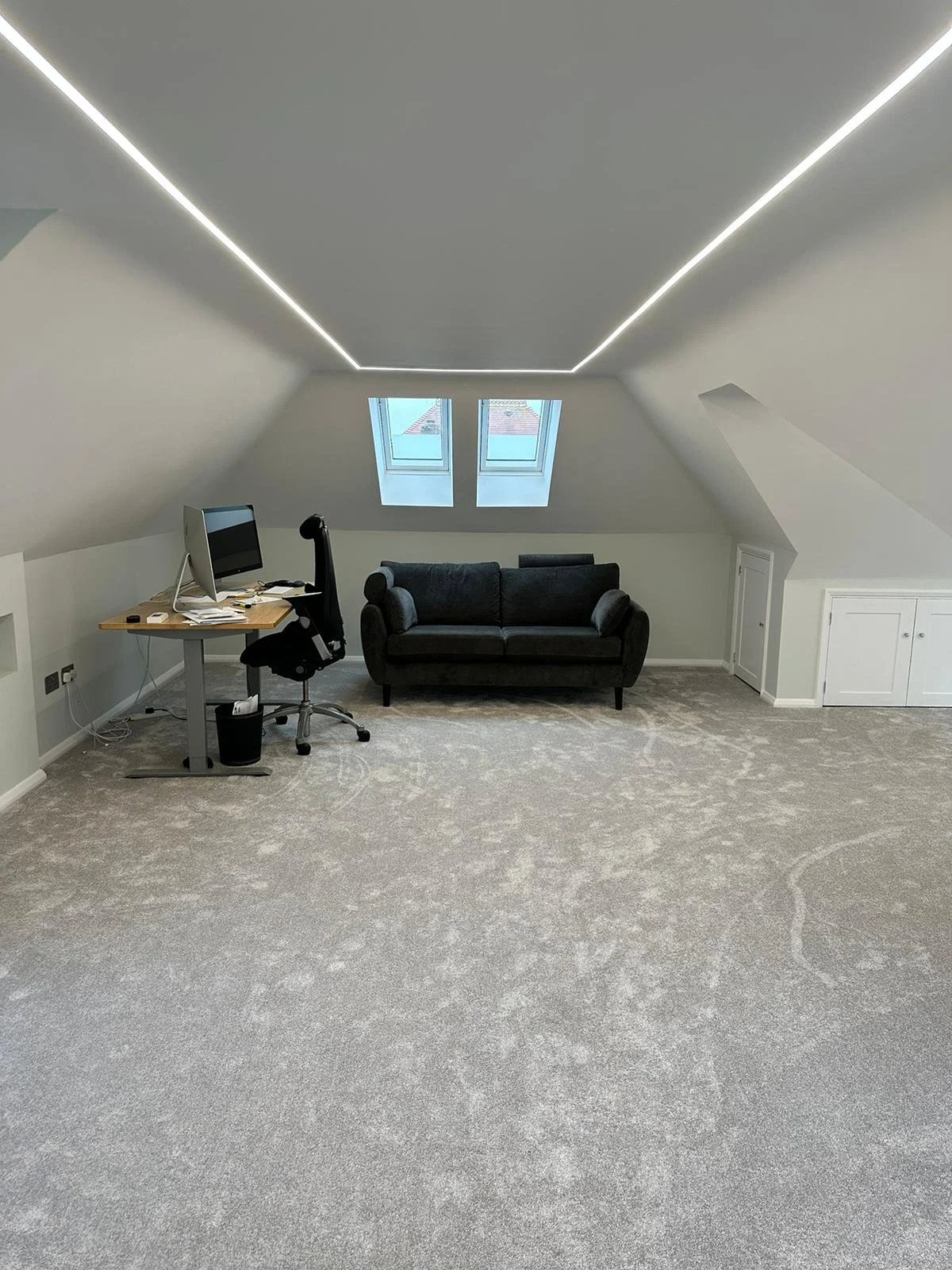 An attic room with sloped ceilings, carpeted floor, a small couch, a desk with a computer, and two windows on the far wall.