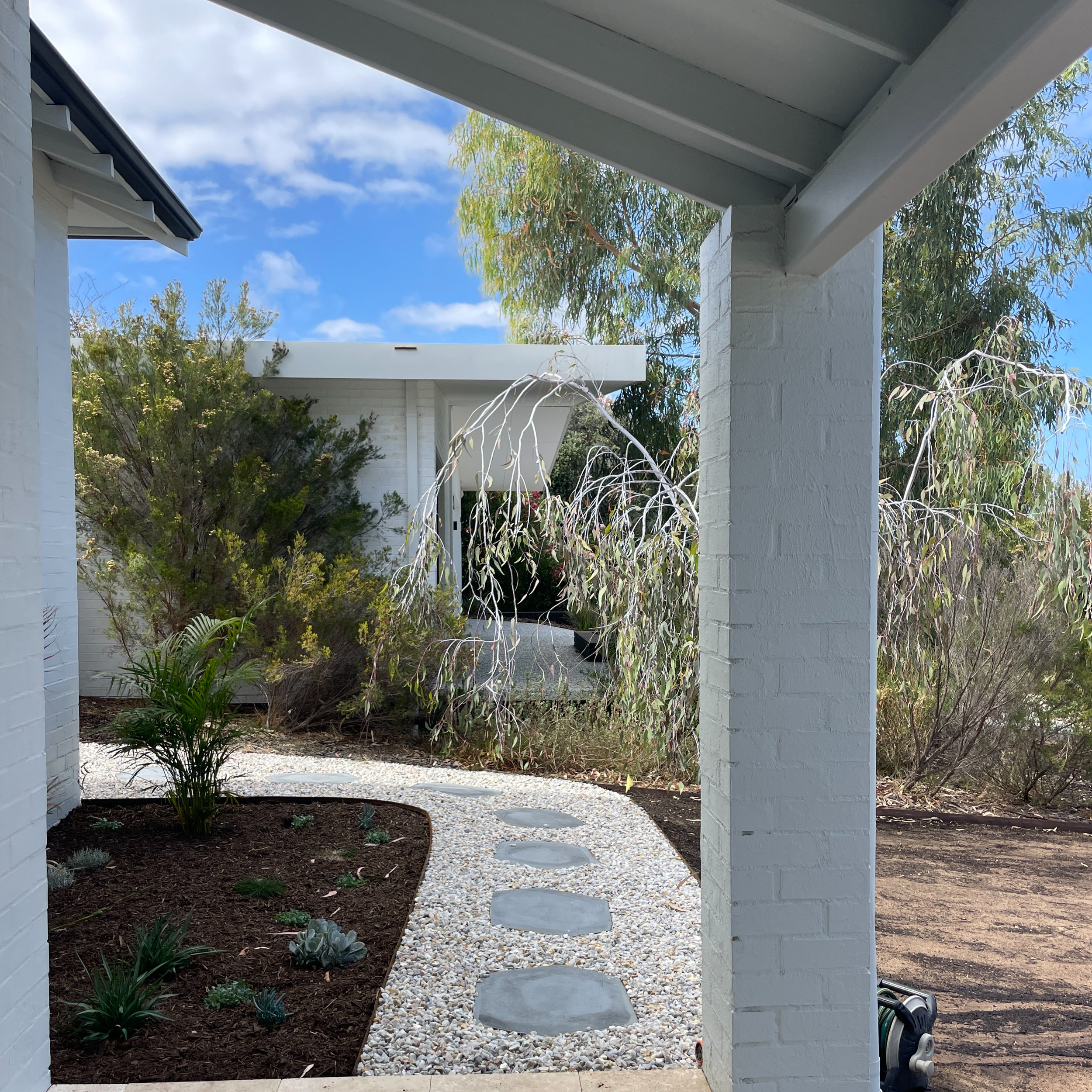View of a white gravel pathway with large stepping stones, surrounded by a landscaped area with plants and shrubs, leading to a white building with a flat roof, under a cloudy sky.
