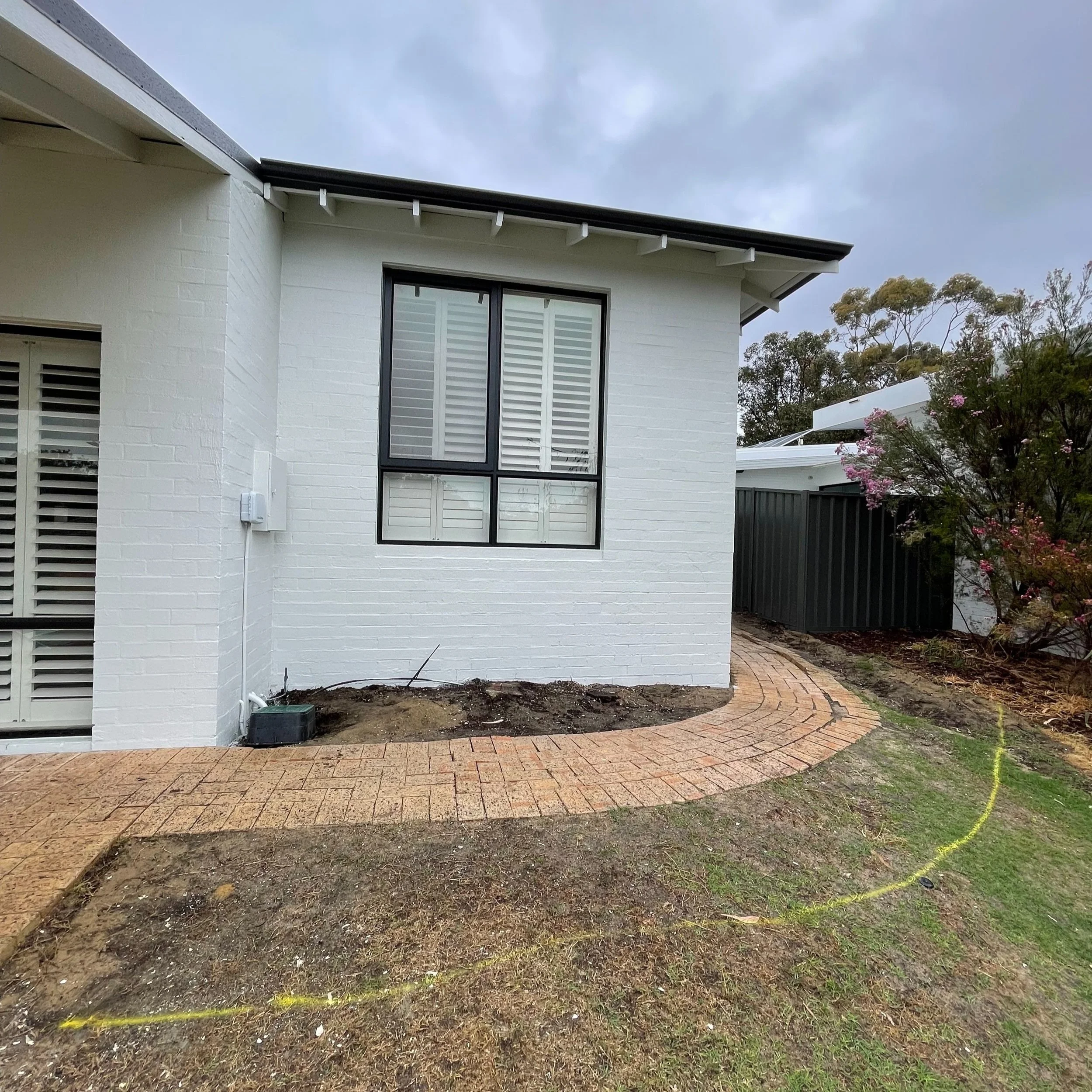 Backyard with brick pathway, dirt patches, a white brick house wall with window, and a tree with pink flowers.