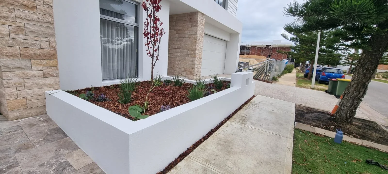A modern house exterior with a white garden bed, small plants, a tree, and a sidewalk in front.