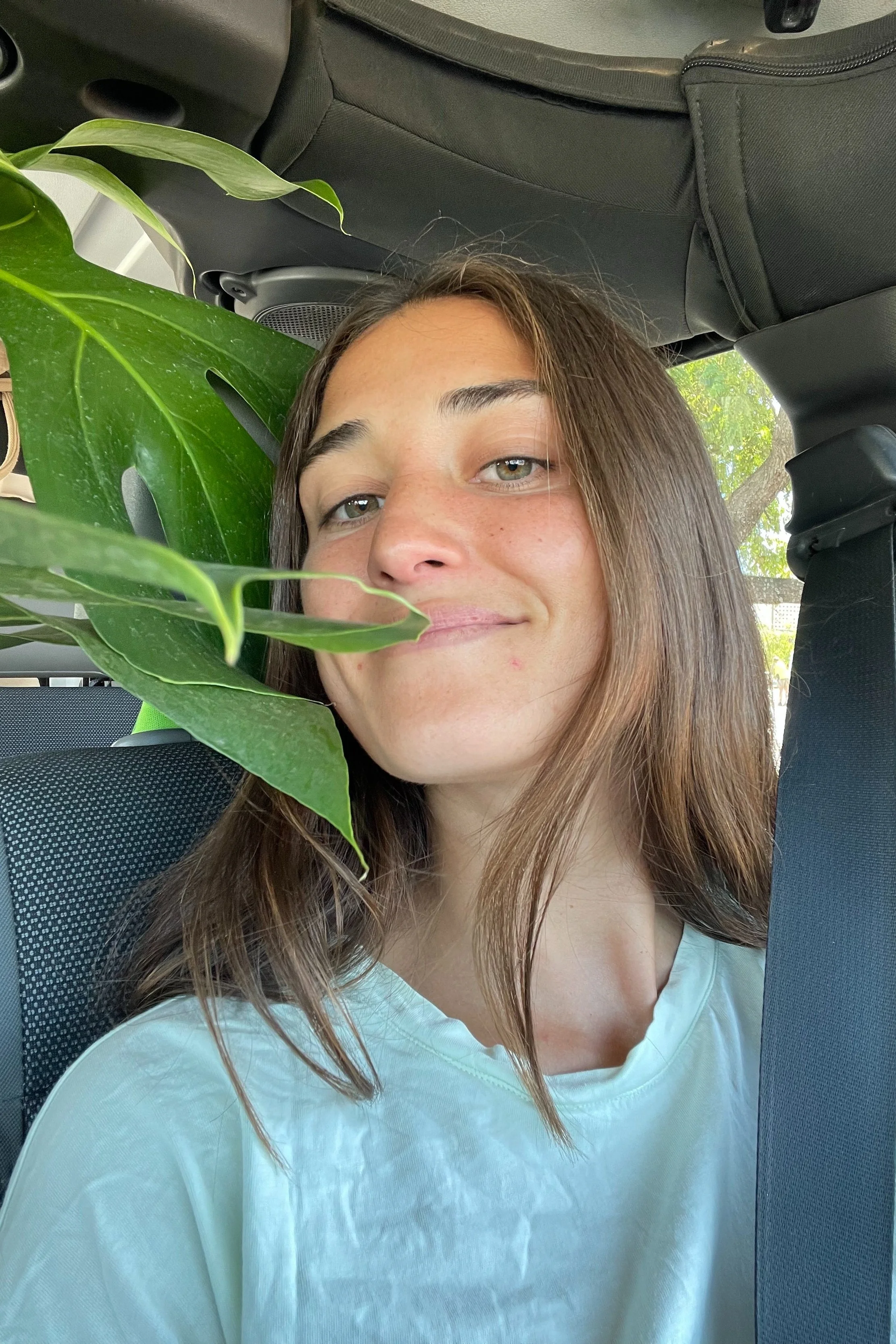 A young woman with light brown hair and fair skin taking a selfie inside a car, with green leaves partially covering her face, and sunlight coming through the window.