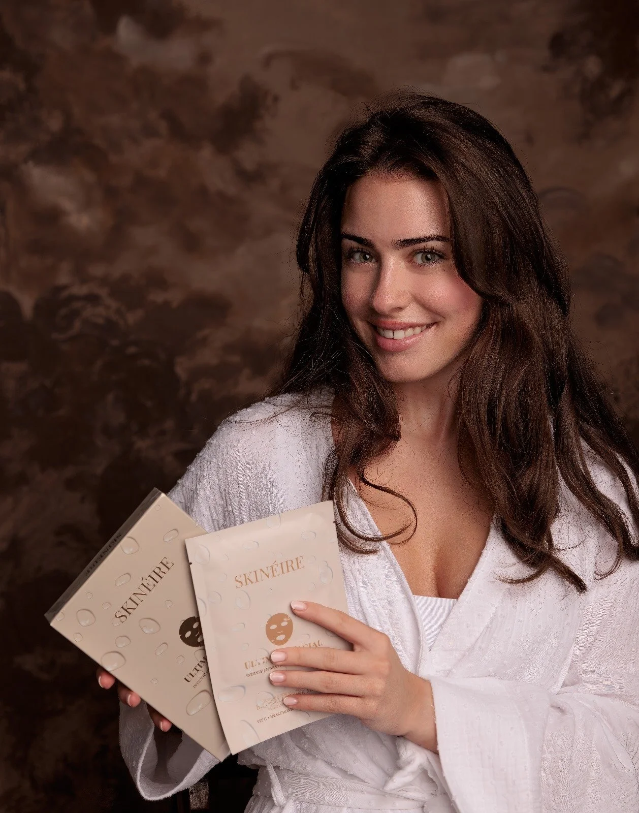 A woman with long brown hair smiling while holding skincare product packages labeled SKINÉIR, dressed in a white robe against a brown textured background.
