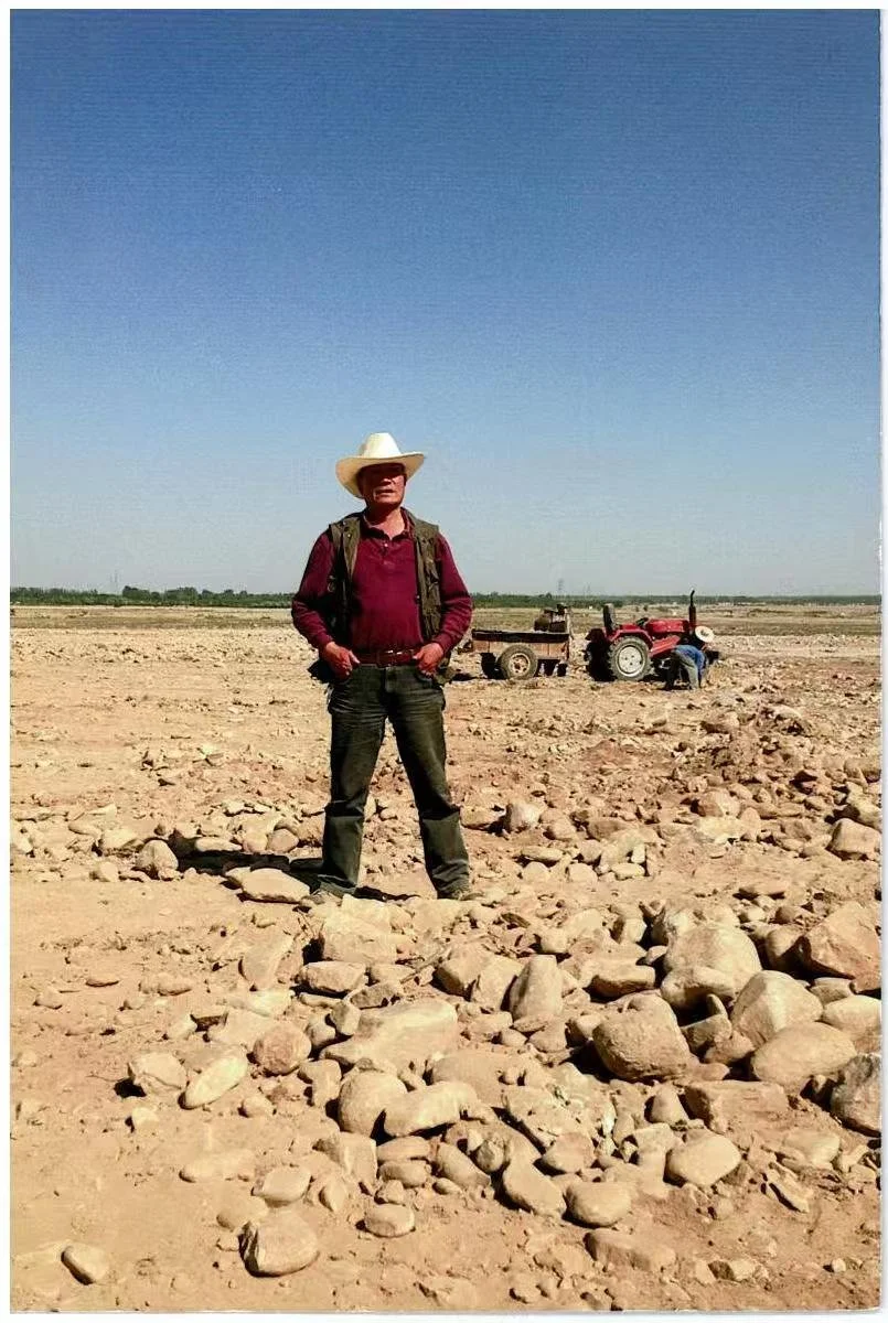 A man standing in a dry, rocky field wearing a cowboy hat, red shirt, and backpack. In the background, there is a tractor and a person working near it on a clear day.
