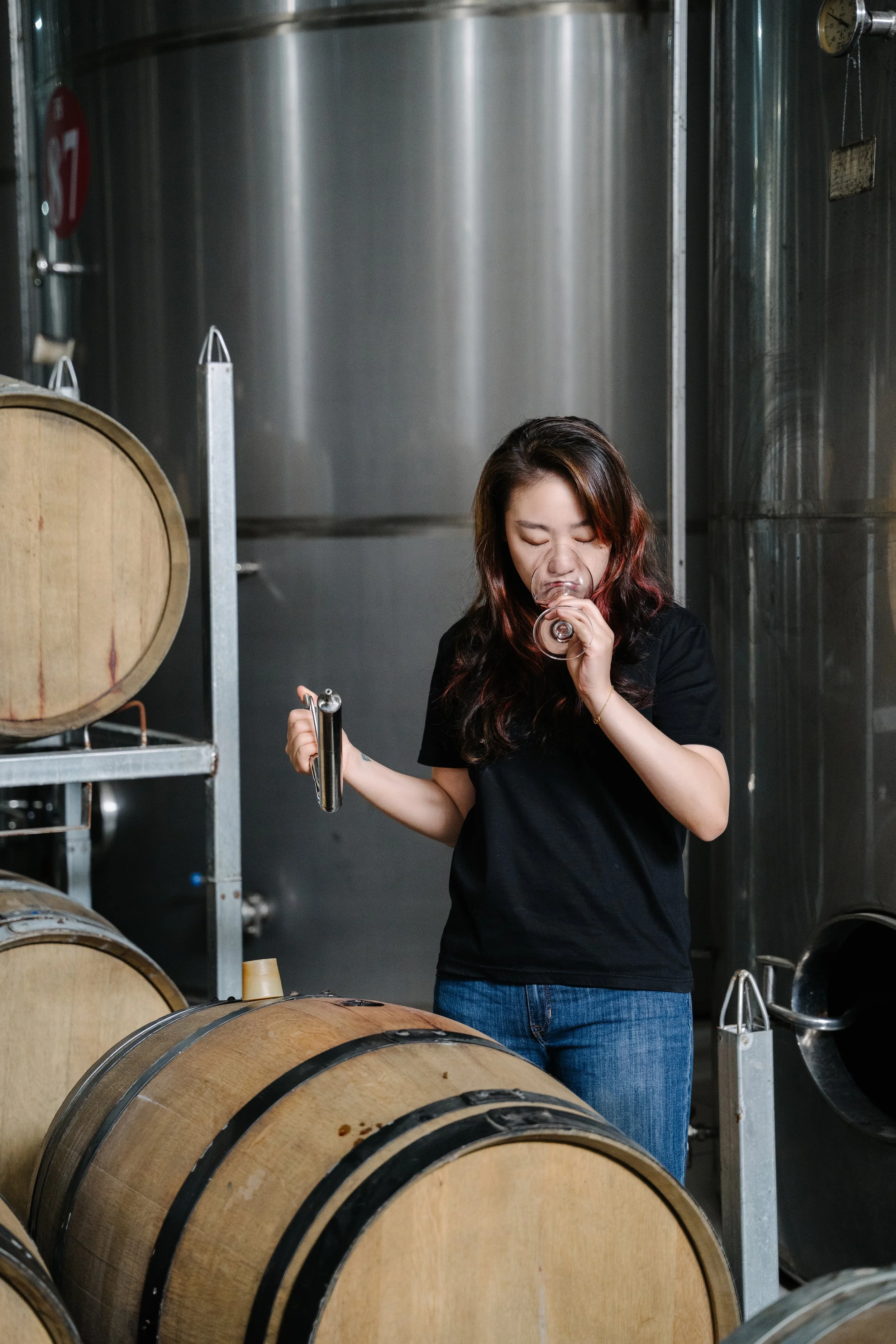 A woman in a wine cellar tasting wine from a glass, holding a wine key, with wooden barrels and stainless steel tanks in the background.