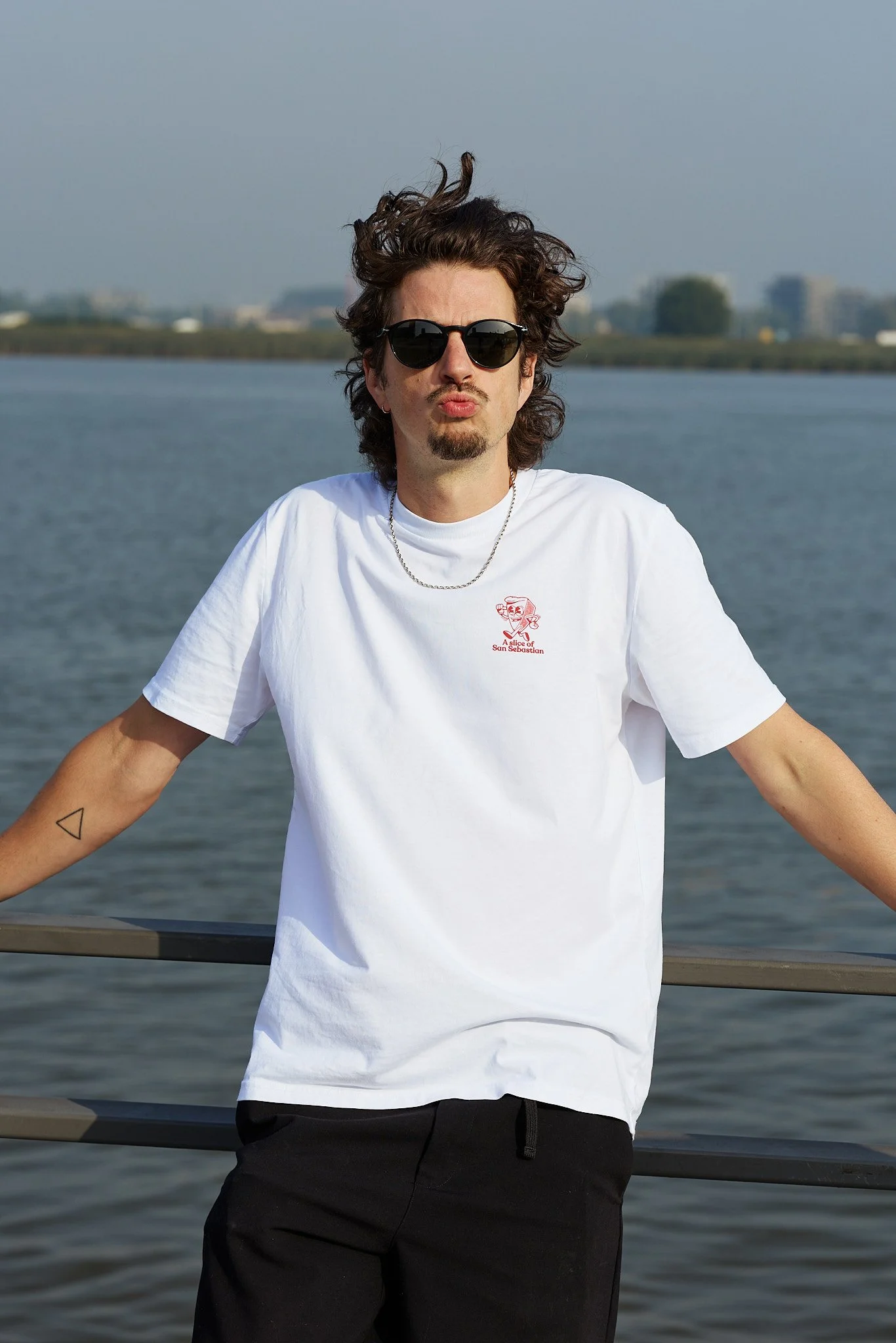 A man with curly hair and sunglasses standing outdoors near water, wearing a white t-shirt and black pants, making a kiss face at the camera.