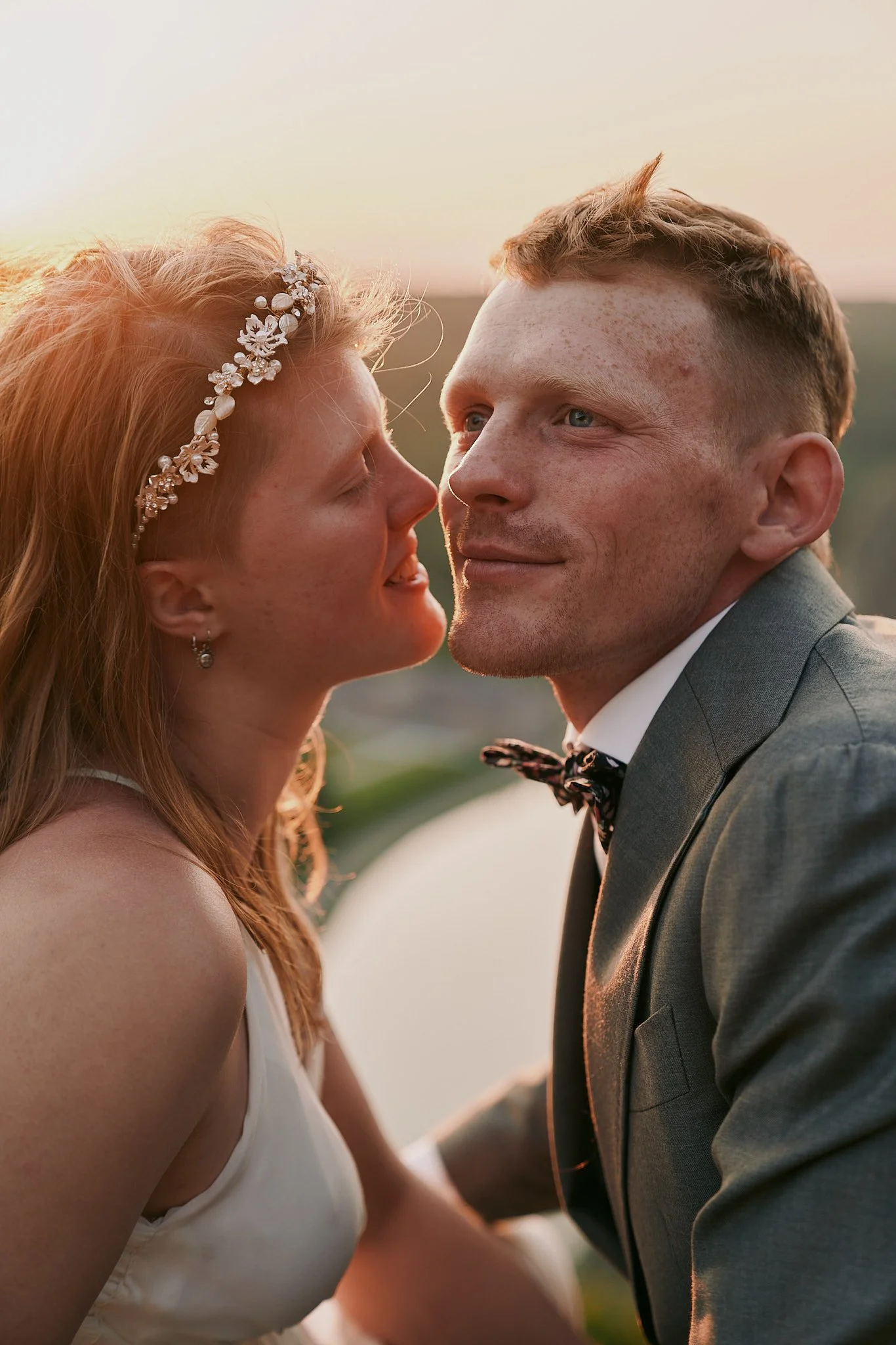 A bride and groom close up smiling at each other outdoors at sunset, with scenic landscape in the background.