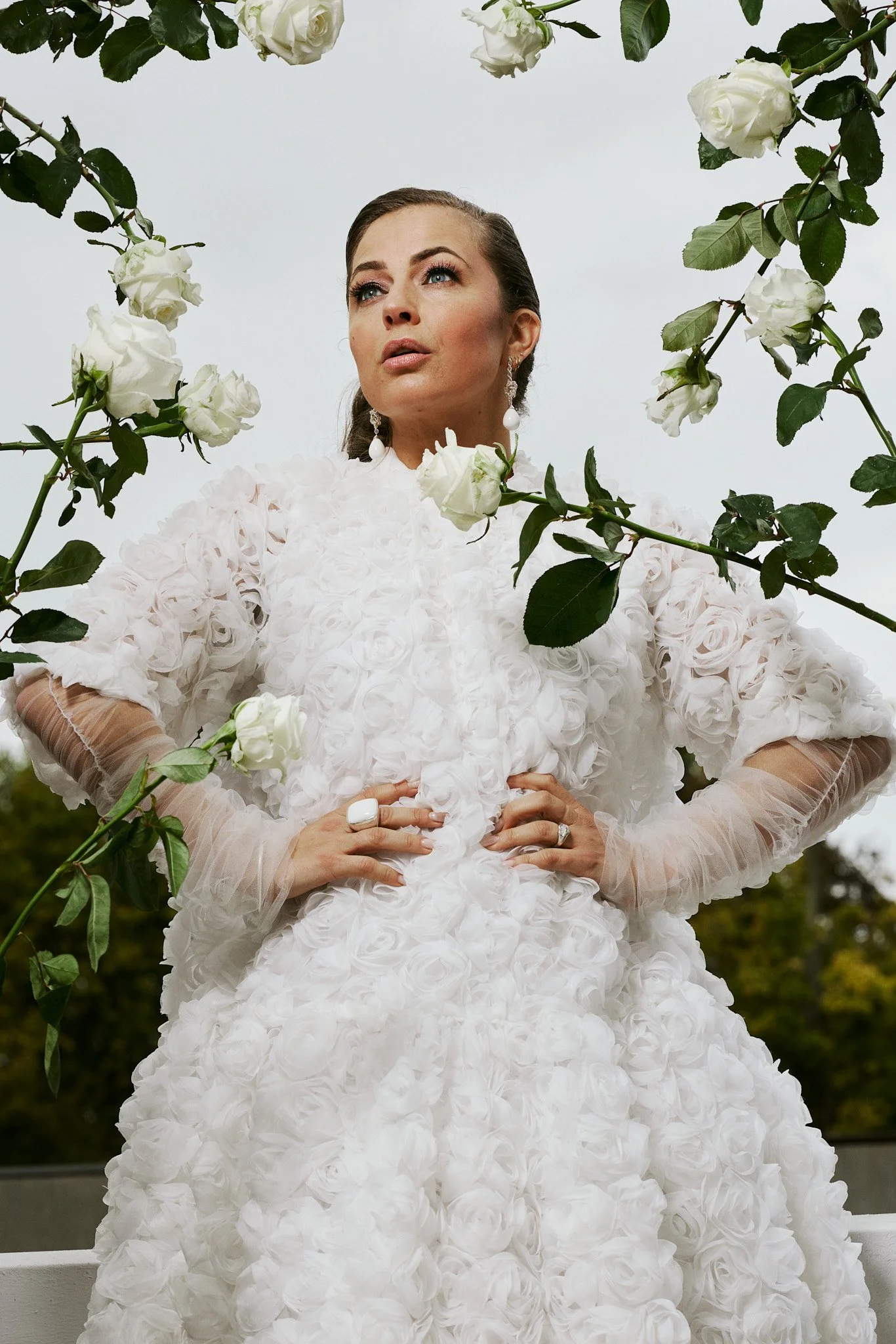 A woman in a white dress adorned with roses, standing among white roses and greenery, with her hands on her hips, looking confidently ahead.