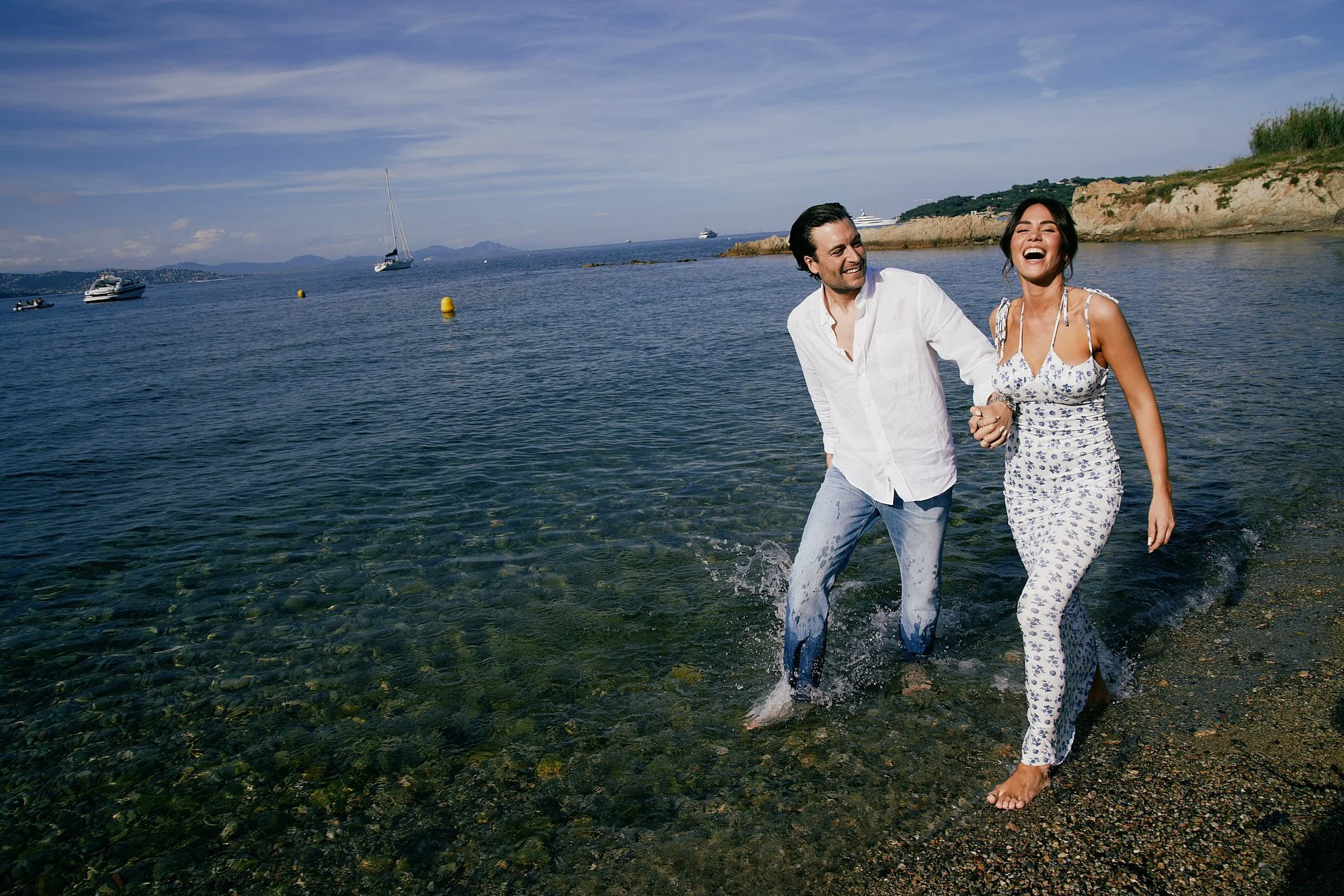 A happy couple walking in the shallow water at the beach, laughing and holding hands, with sailboats and yachts on the water and a rocky shoreline in the background.