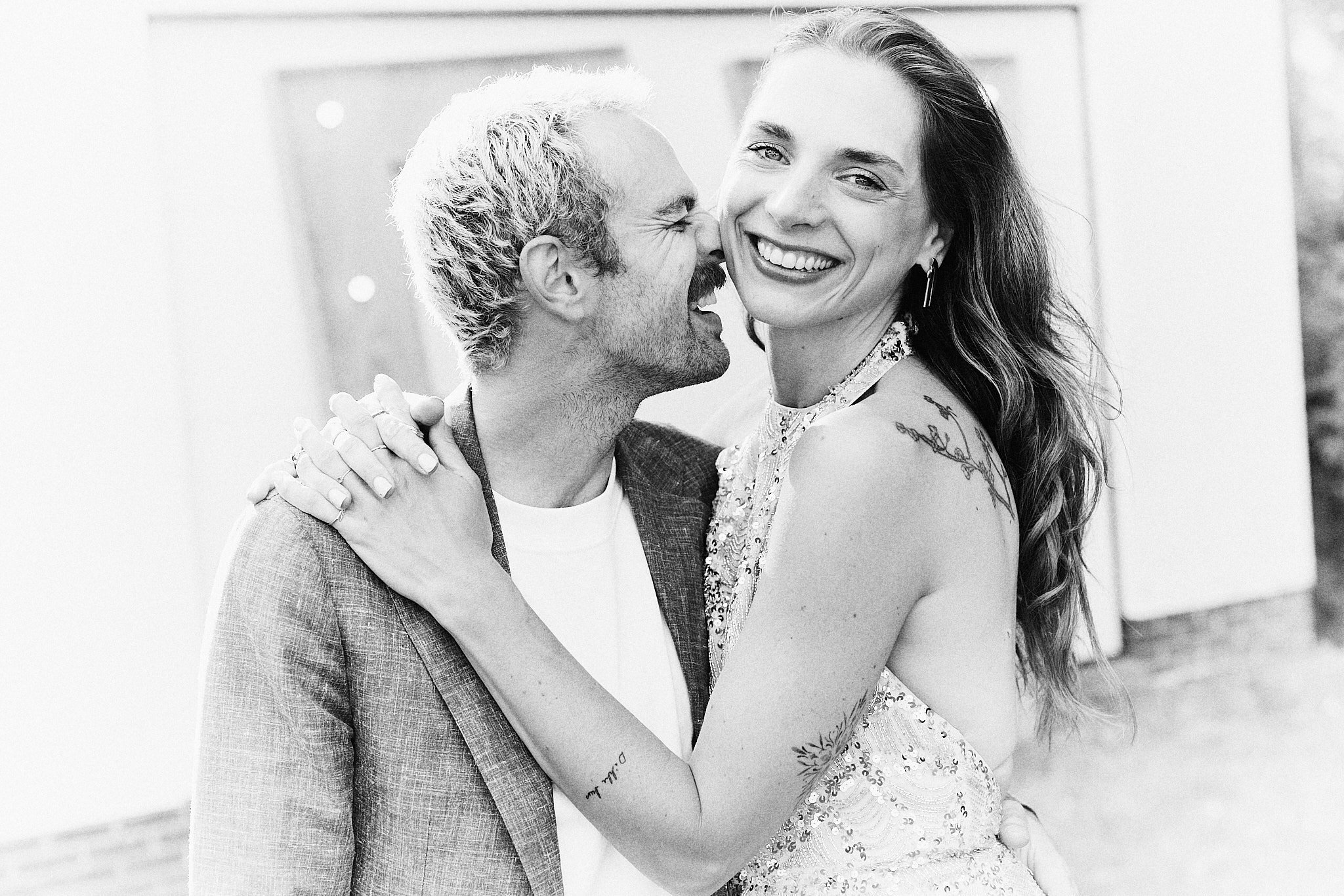 A joyful man with light hair and beard in a suit embraces a smiling woman with long hair in a sleeveless dress, sharing a hug, outdoors, black and white photo.