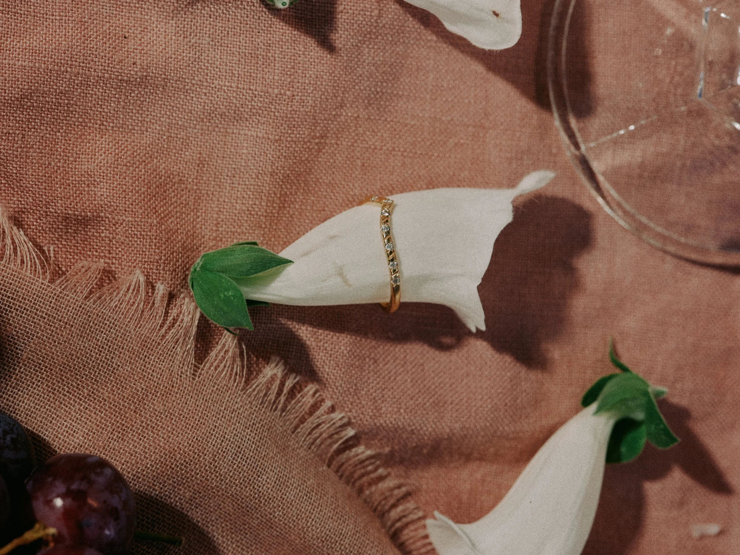 A white flower with green leaves on a brown fabric surface, with a gold ring with small gemstones placed on the flower. Part of a glass is visible on the top right corner.