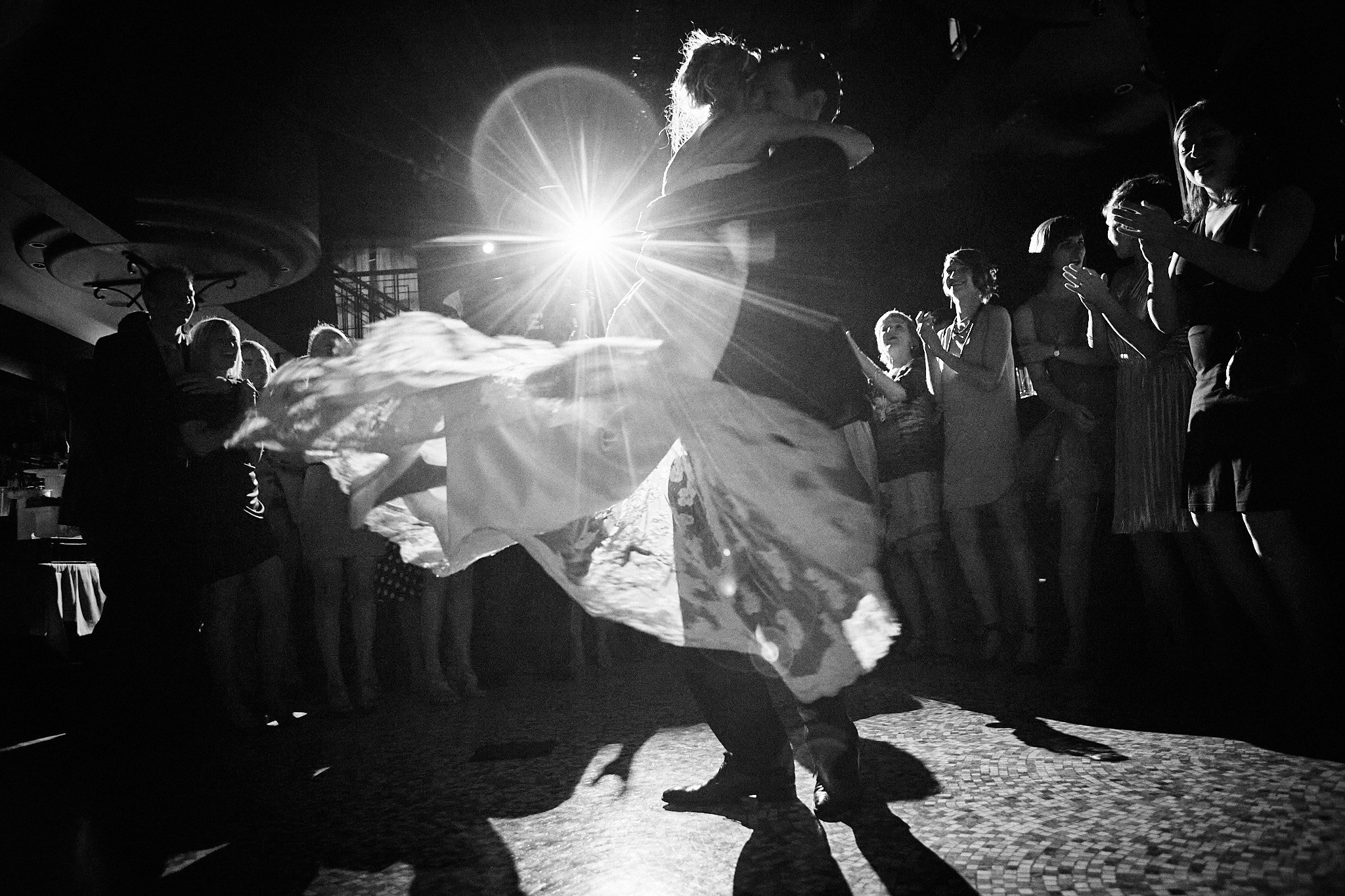 Couple dancing at a wedding reception with guests clapping and watching, silhouetted against a bright light.