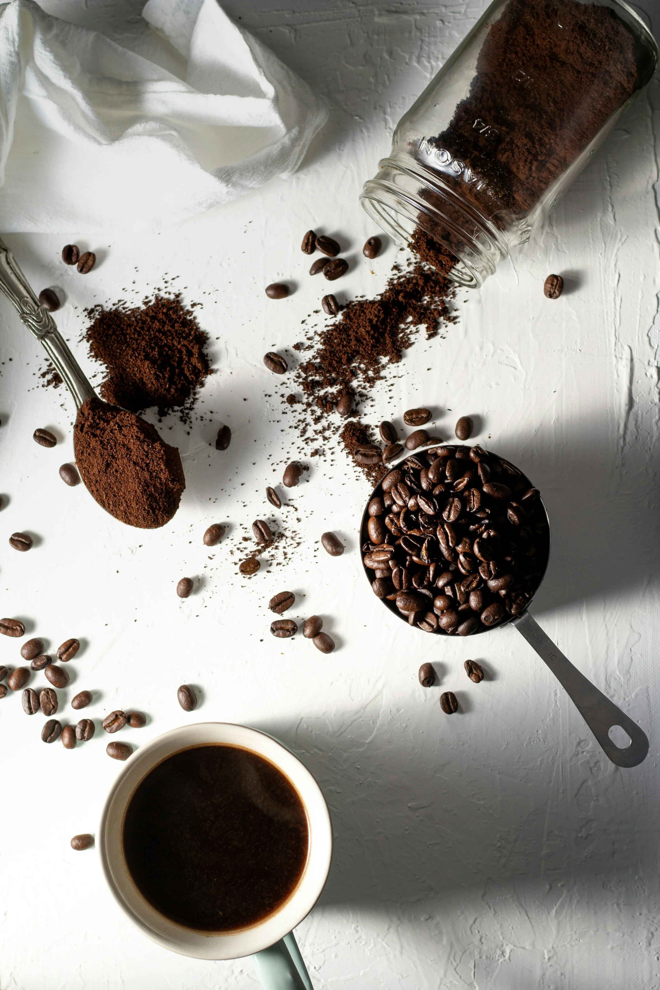 Coffee beans spilled from a jar, ground coffee on a spoon, and a cup of brewed coffee on a white textured surface.