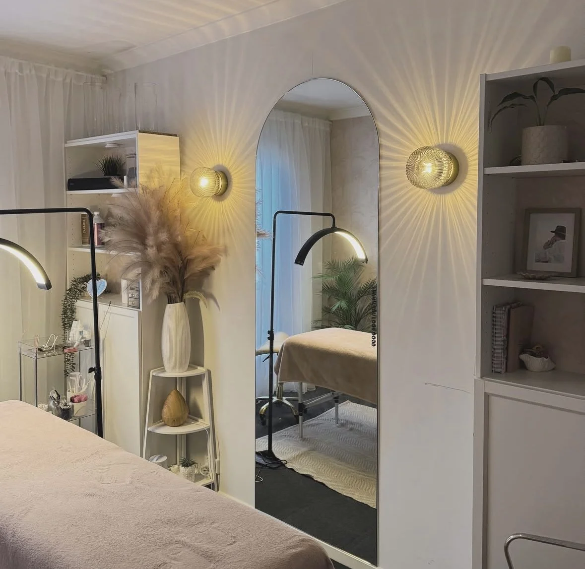 A cozy room with a large mirror, wall sconces, a bed, and decorative plants.