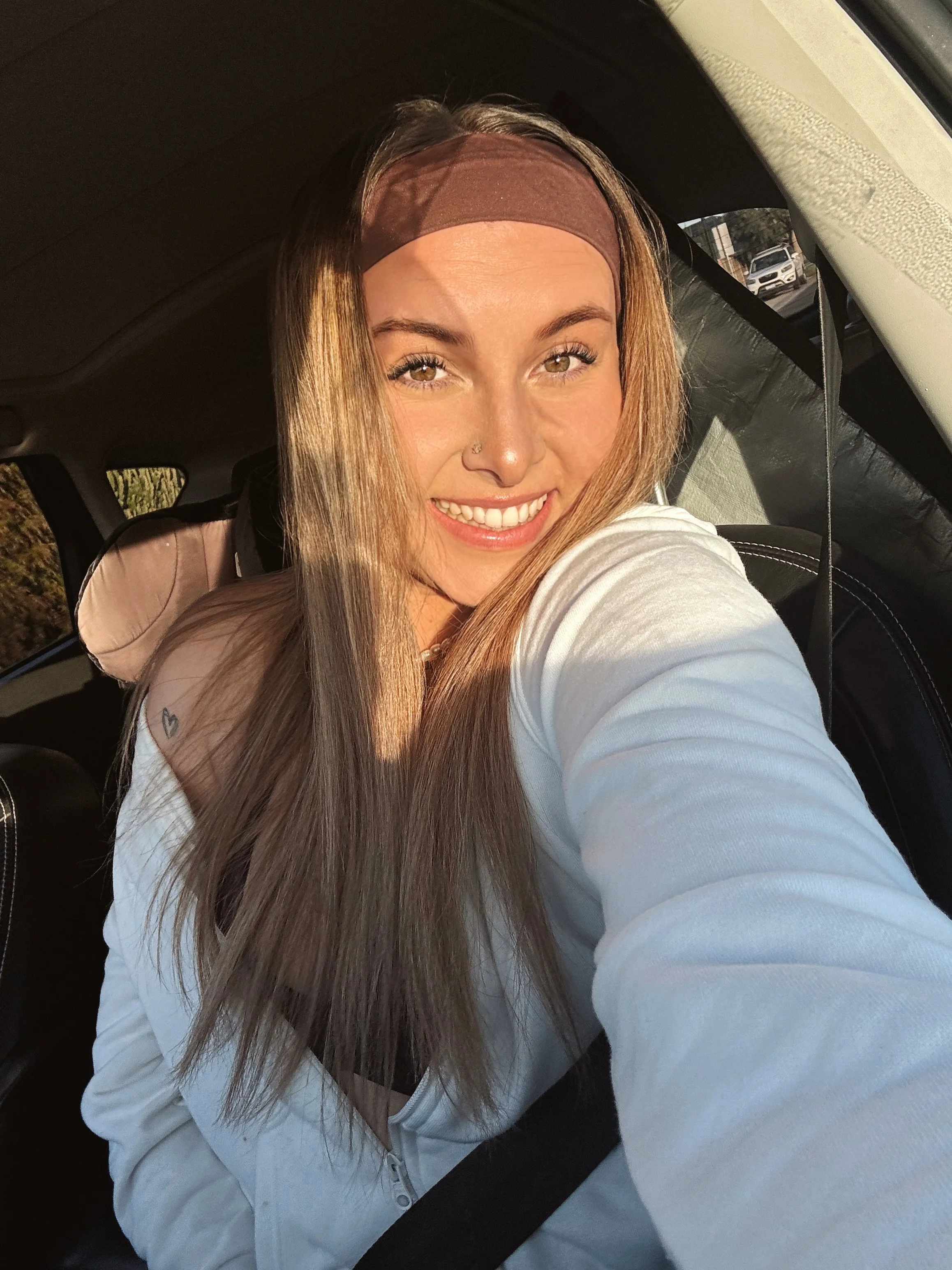 A woman with long brown hair, wearing a brown headband and white jacket, taking a selfie inside a car with sunlight on her face and a bright smile.