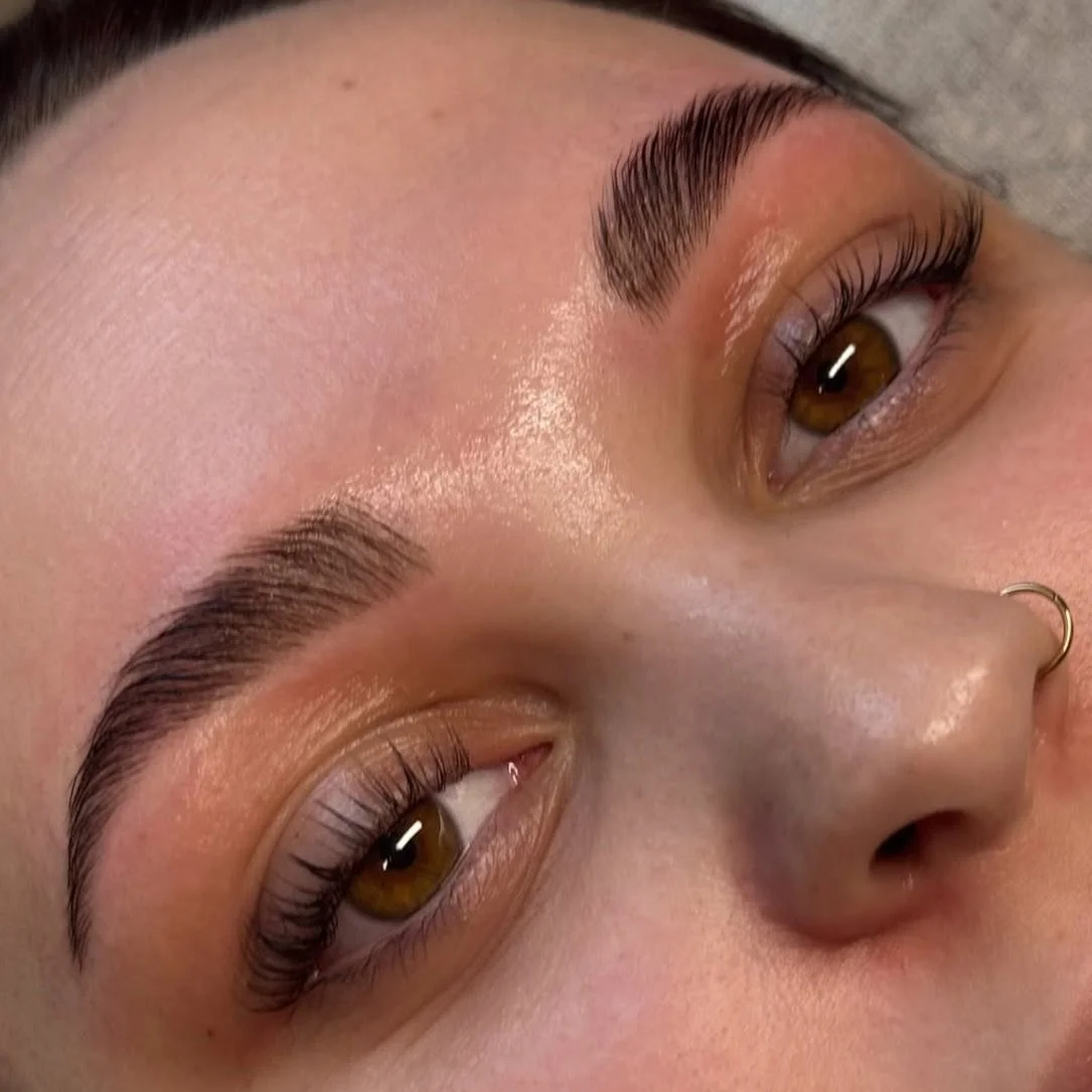 Close-up of a person's face showing well-groomed eyebrows, brown eyes, and a nose with a small hoop nose ring, with smooth skin and natural makeup.