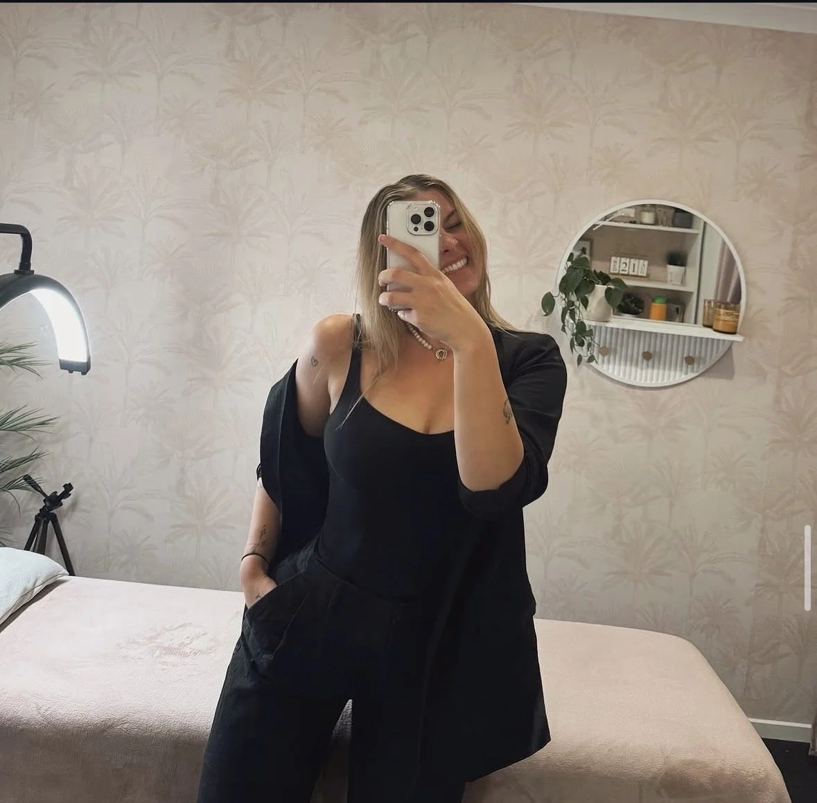A woman taking a mirror selfie in a room with light patterned wallpaper. She is smiling, wearing a black top and black pants, with a black jacket draped over her shoulders. There is a bed with a light-colored blanket, a round mirror on the wall, and a shelf with plants and decor in the background.