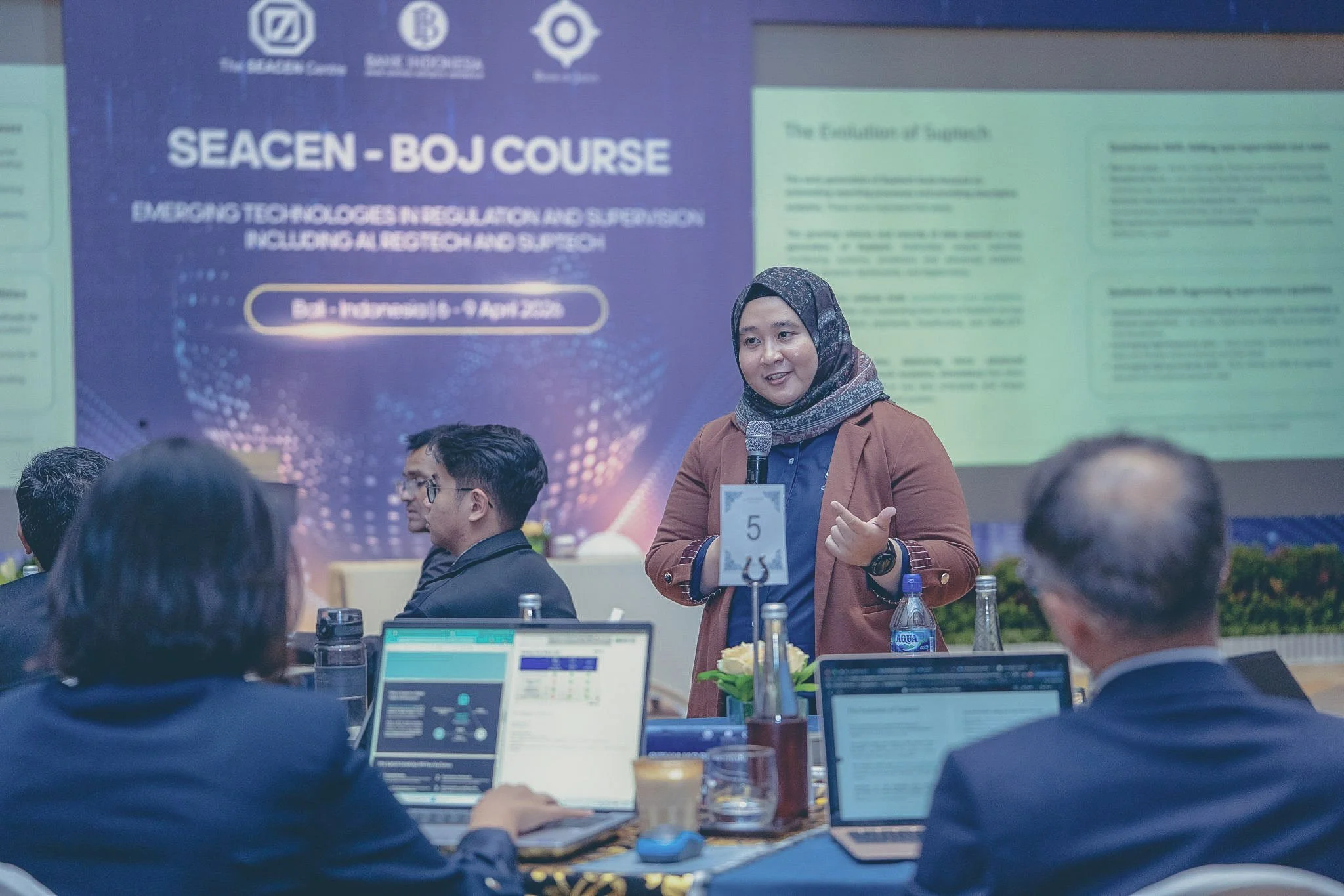 Amanah Ramadiah stands with a microphone in front of a presentation, surrounded by seated people, several laptops, and conference materials. The background shows a large blue and green banner with conference information.