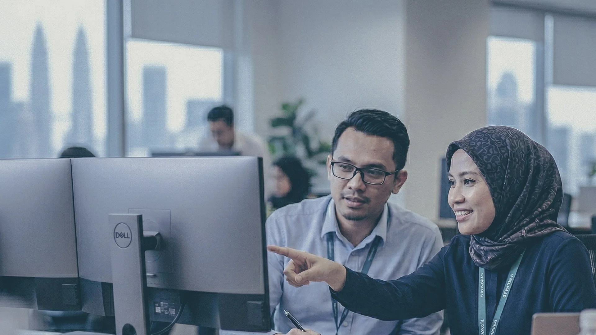 Two coworkers, a man and a woman, discussing work at a computer in an office environment. The woman is pointing at the monitor while the man is looking on attentively. Other employees are visible working in the background.