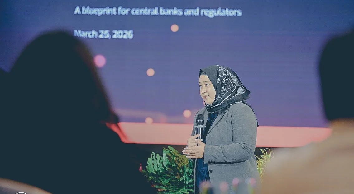 FNA's Amanah Ramadia speaking into a microphone at a conference. A large screen behind her displays the text 'A blueprint for central banks and regulators, March 25, 2026,' with a dark background and colored dots.