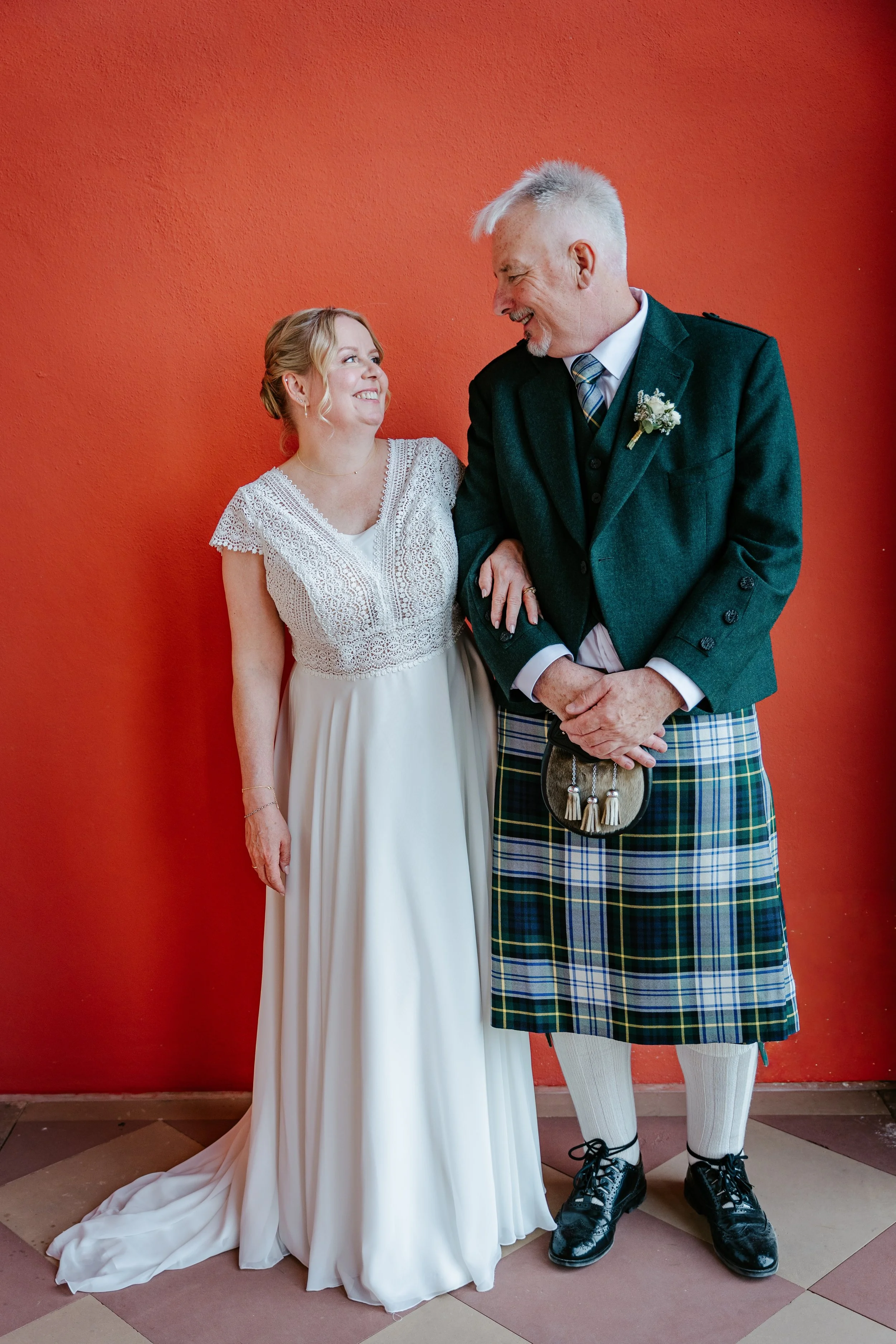 Lachendes Brautpaar vor roter Wand: Braut im weißen Spitzenkleid und Bräutigam im grünen Kilt mit Blumenanstecker.