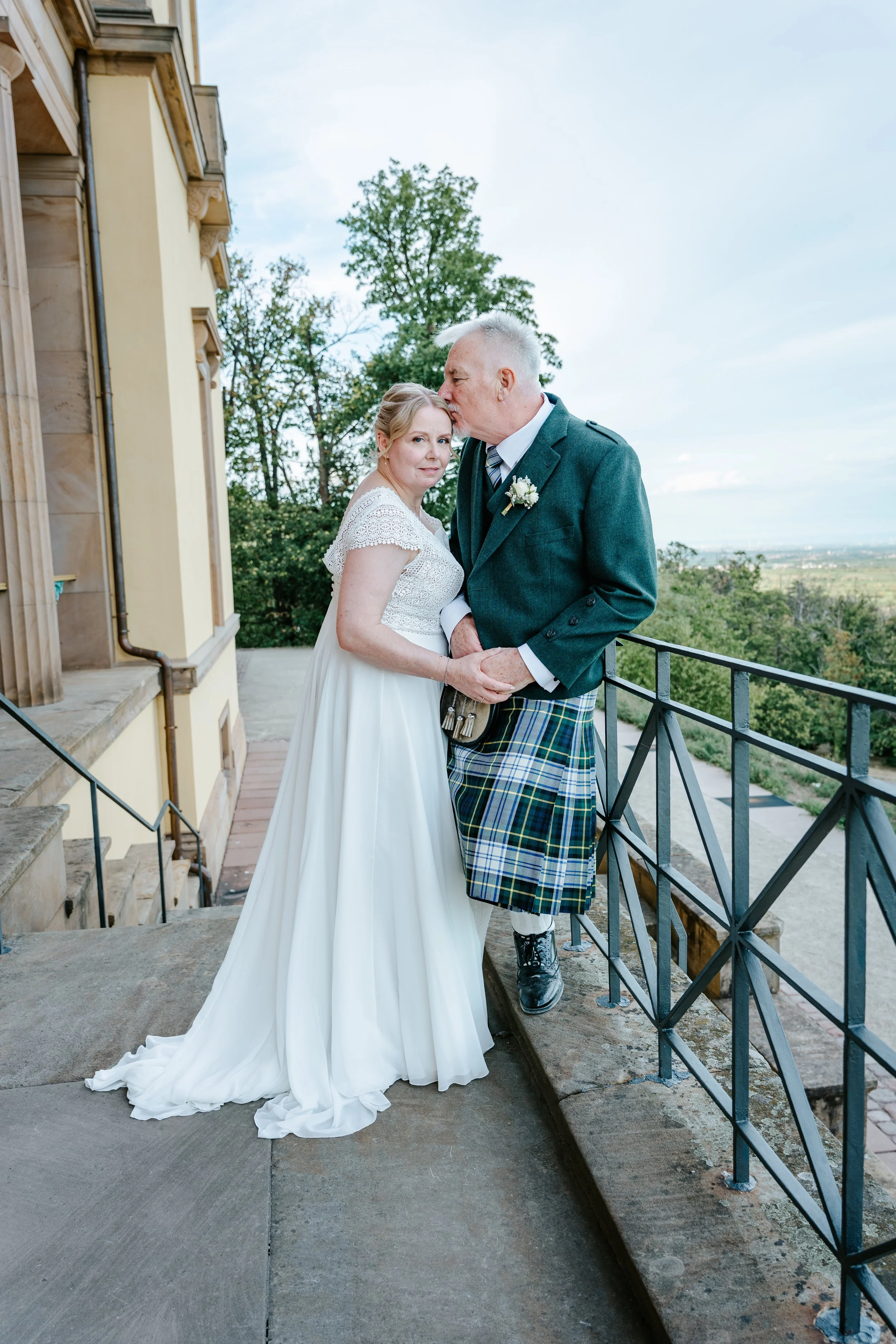 Brautpaar auf einer Terrasse: Der Bräutigam im grünen Kilt küsst die Braut im weißen Hochzeitskleid vor einer historischen Villa.