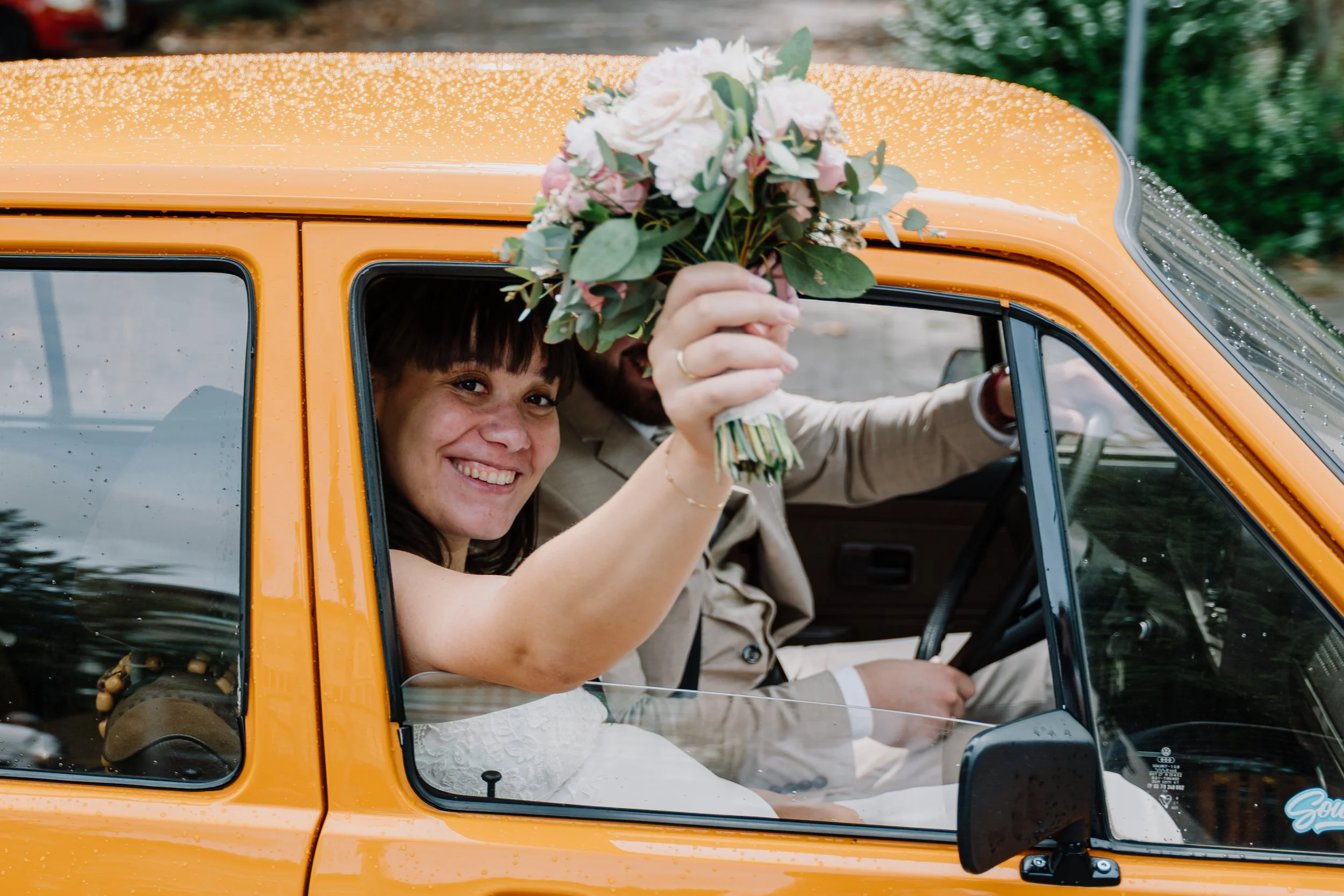 Lächelnde Braut mit Blumenstrauß neben dem Bräutigam in einem orangefarbenen Oldtimer