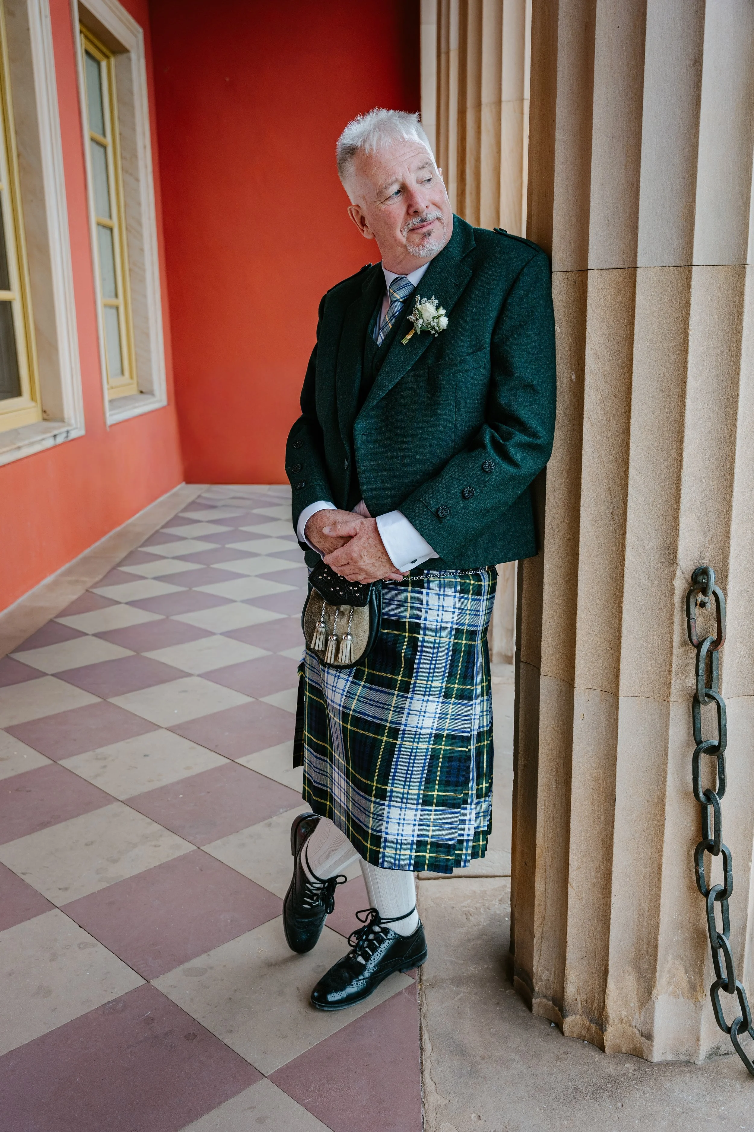 Mann im grünen Kilt mit Ansteckblume, Weste und Krawatte steht in einem historischen Gebäude.