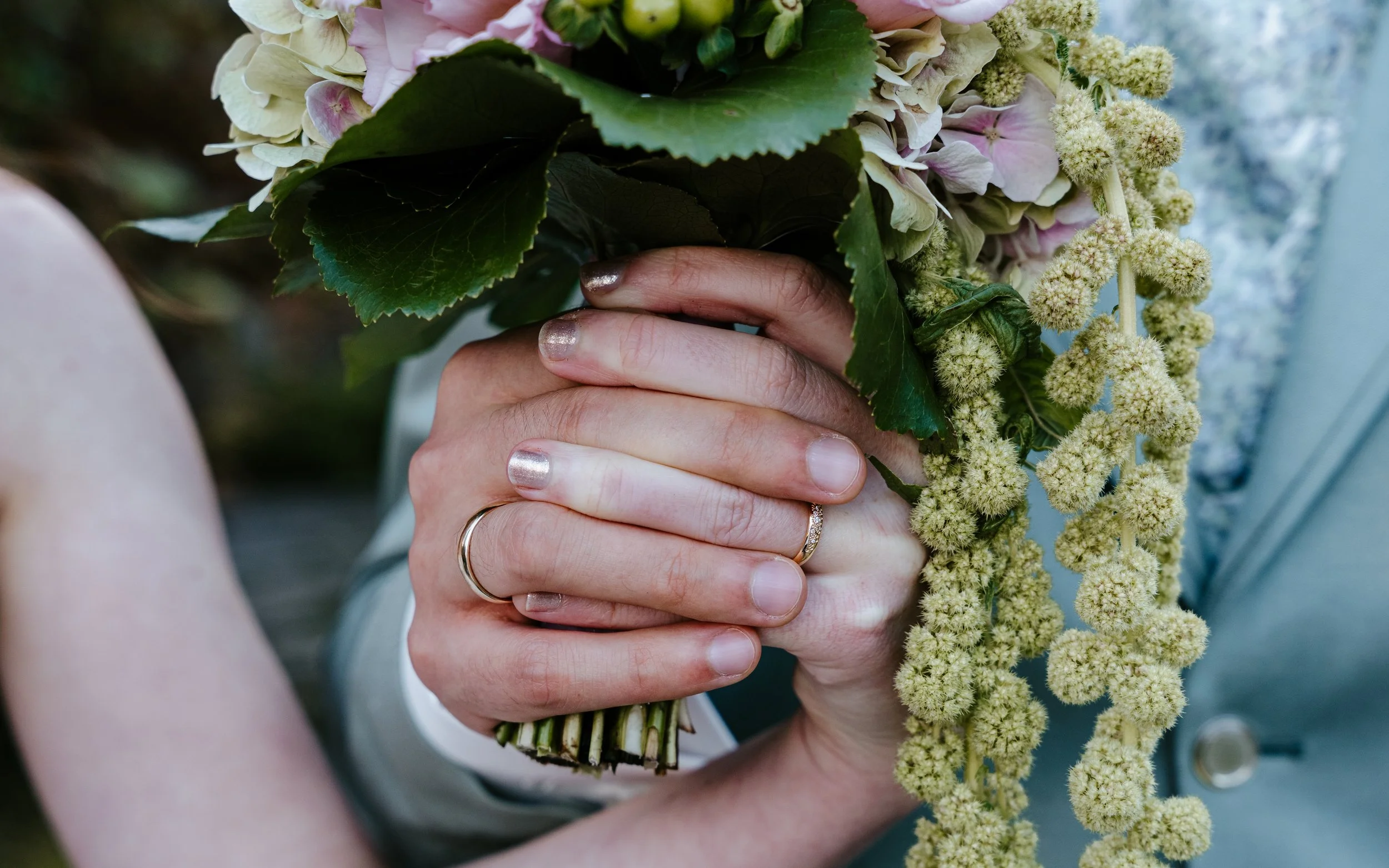 Nahaufnahme einer Person mit Ringen, die einen Blumenstrauß aus rosa-weißen Blüten und grünen Blättern hält