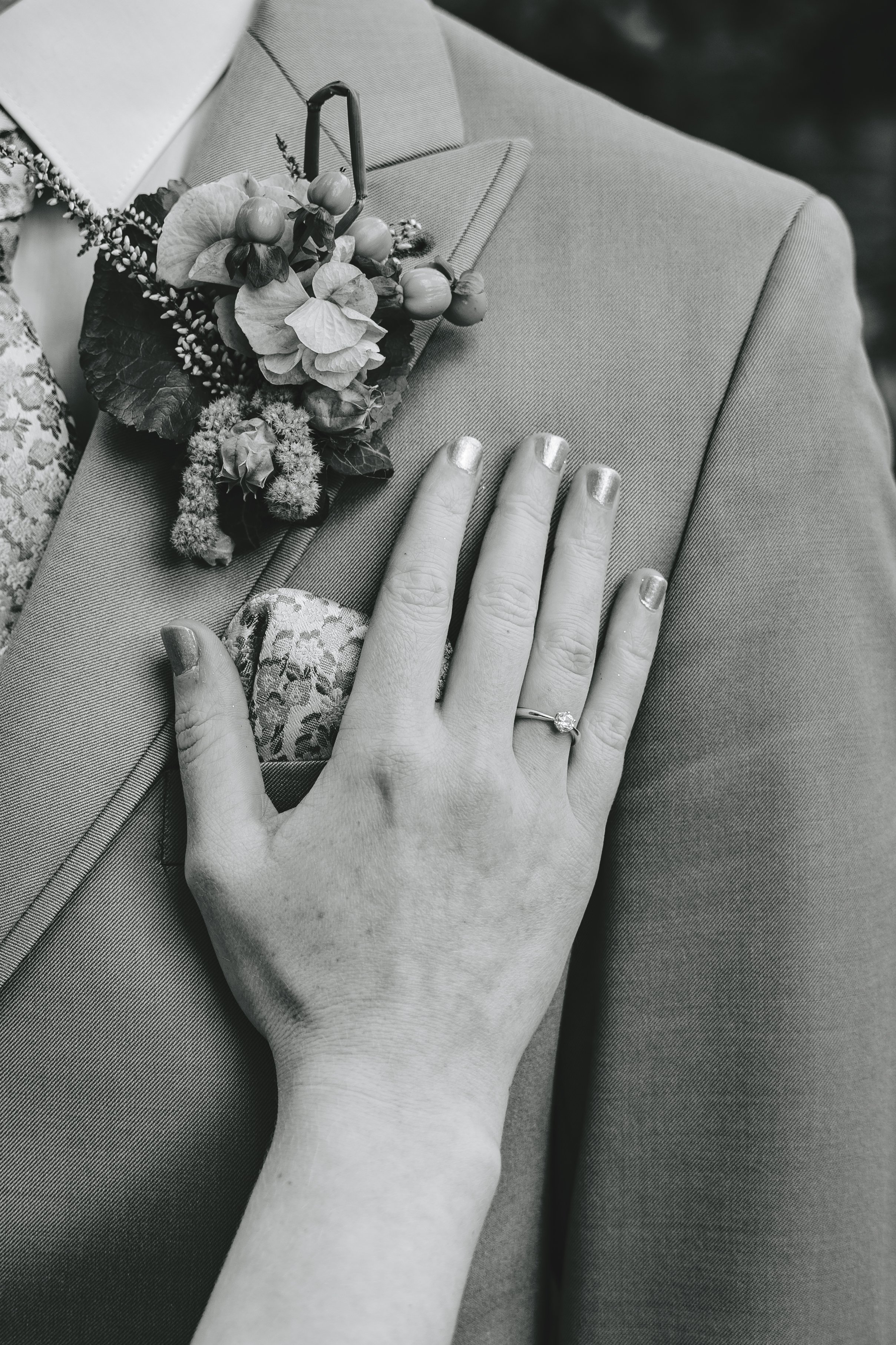Nahaufnahme einer Hand mit silbernem Ring und metallisch lackierten Nägeln auf einem Blazer neben einem floralen Ansteckstrauß.