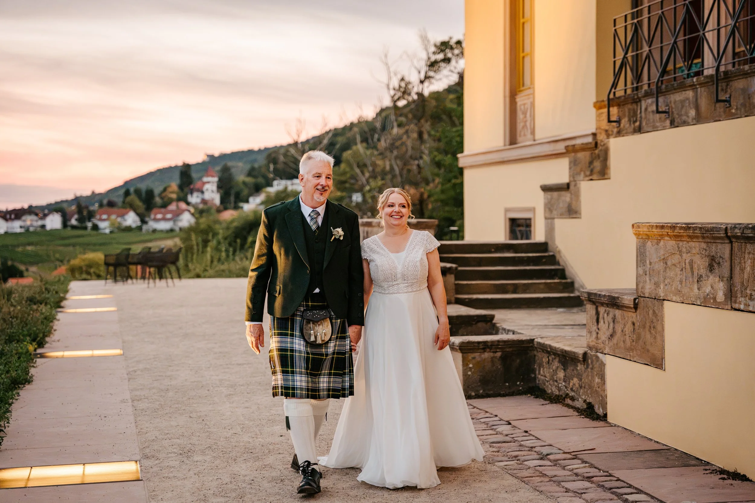Brautpaar vor historischem Gebäude bei Sonnenuntergang: Die Braut trägt ein weißes Kleid, der Bräutigam einen Kilt.