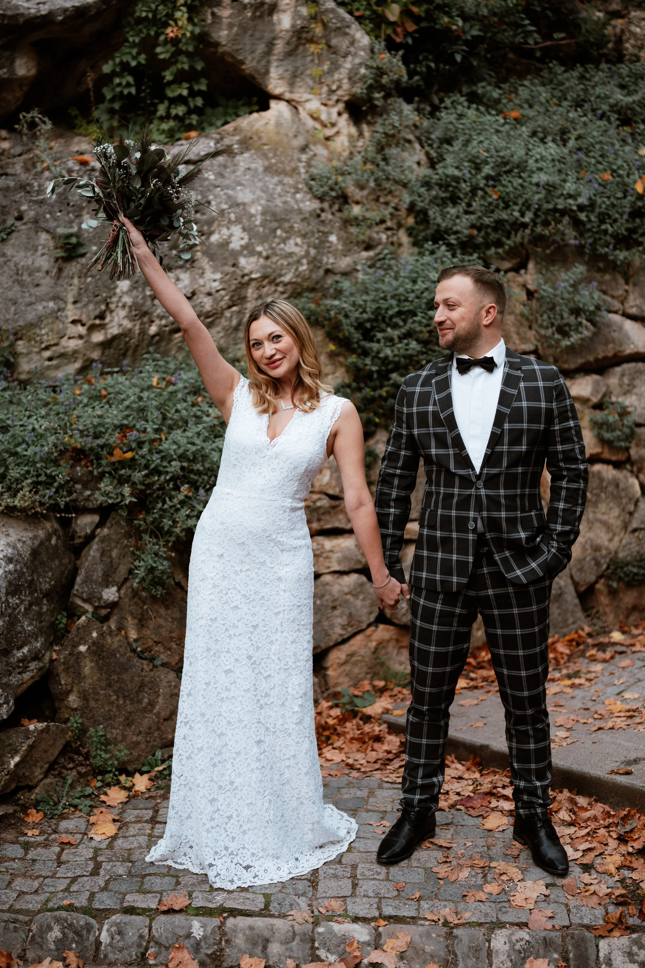 Ein Brautpaar hält Händchen, die Frau trägt ein weißes Brautkleid und hält einen Blumenstrauß hoch, der Mann trägt einen schwarzen Anzug mit kariertem Muster und Fliege, im Hintergrund Steine und Herbstlaub.