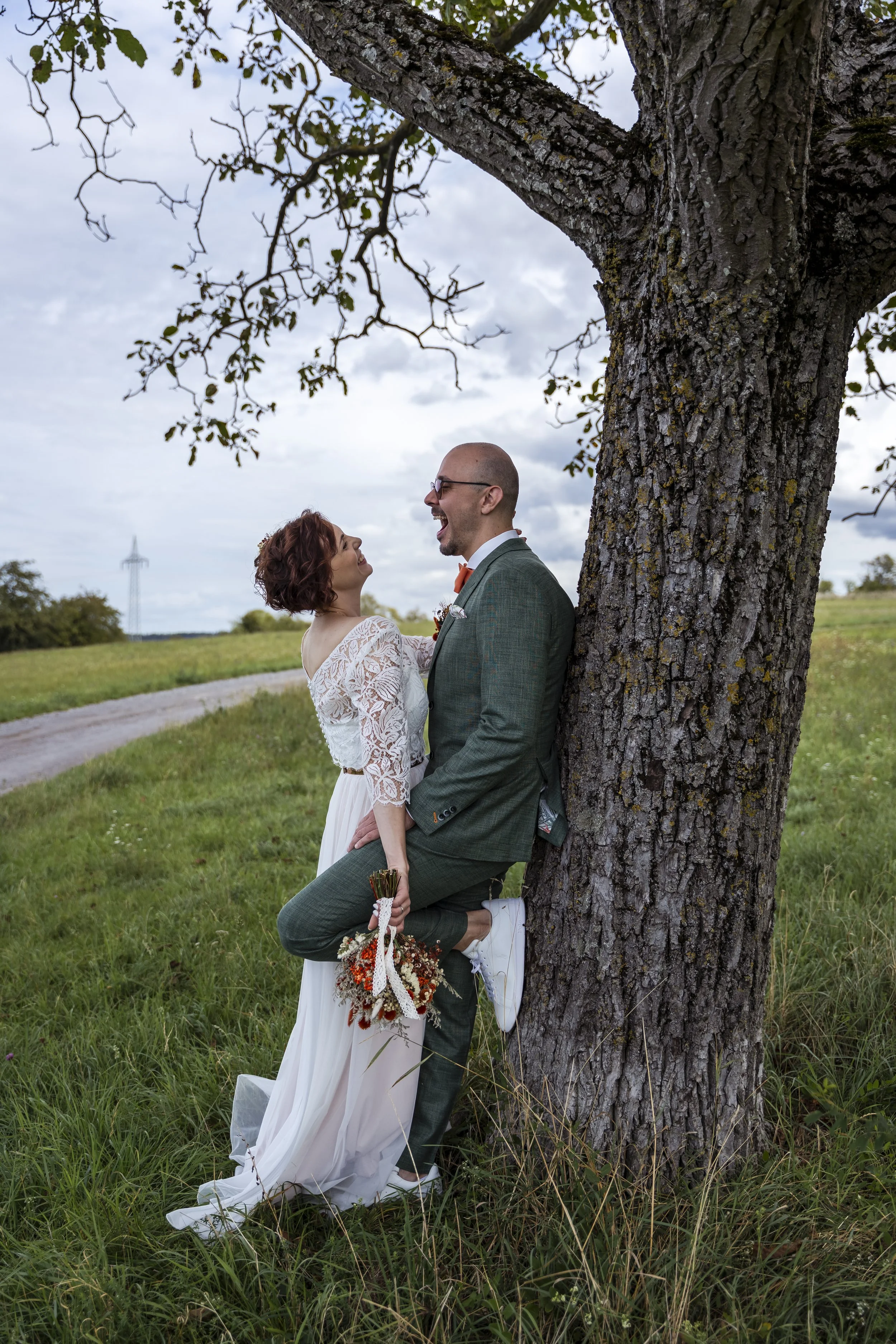 Ein Paar im Hochzeitskleid und Anzug steht unter einem Baum in einer grünen Landschaft. Sie lachen und schauen sich liebevoll an.