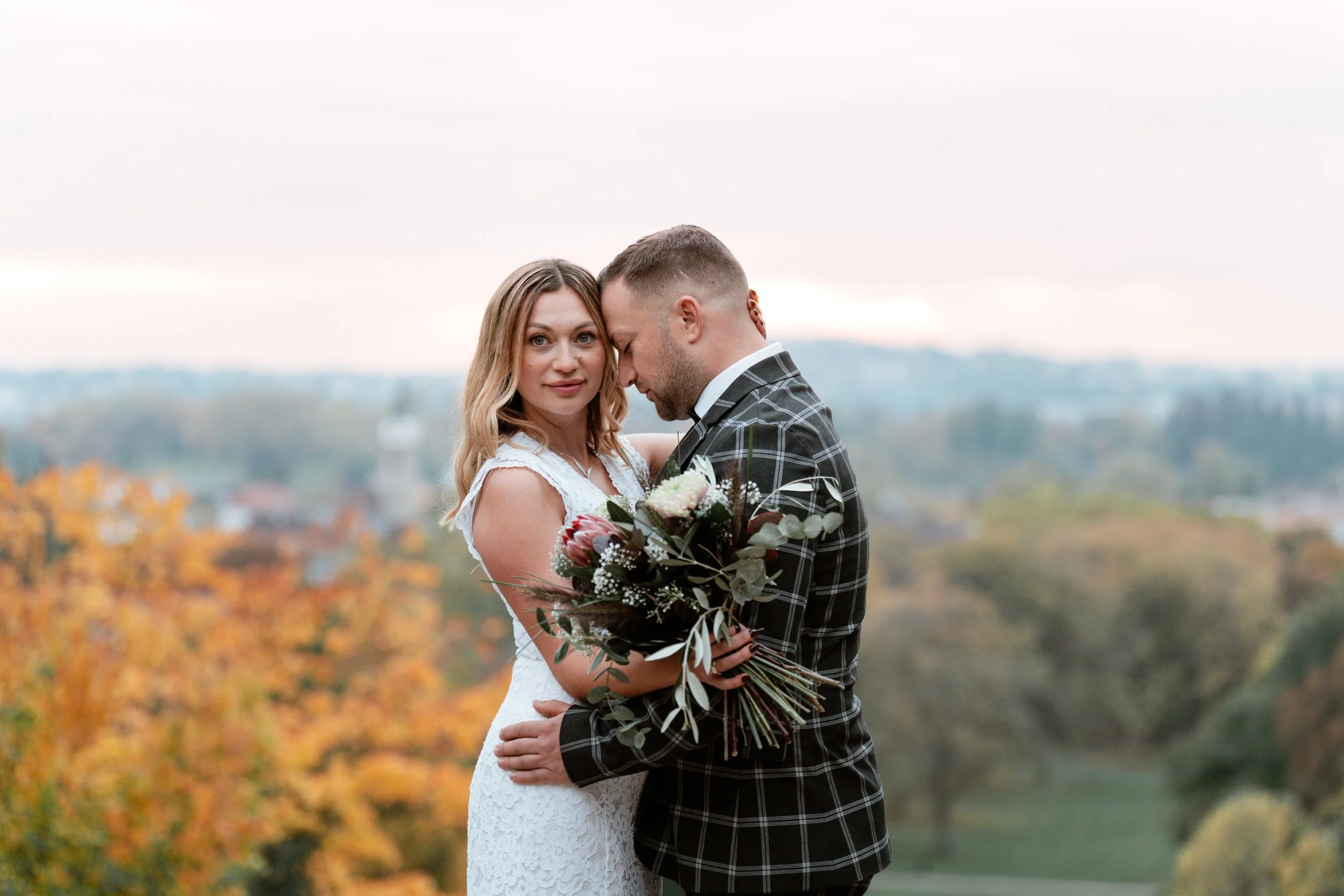 Ein Brautpaar im Freien mit herbstlicher Landschaft, die Frau trägt ein weißes Hochzeitskleid und hält einen Blumenstrauß, der Mann trägt einen schwarzen Anzug mit Karo-Muster, sie posieren eng umarmt.