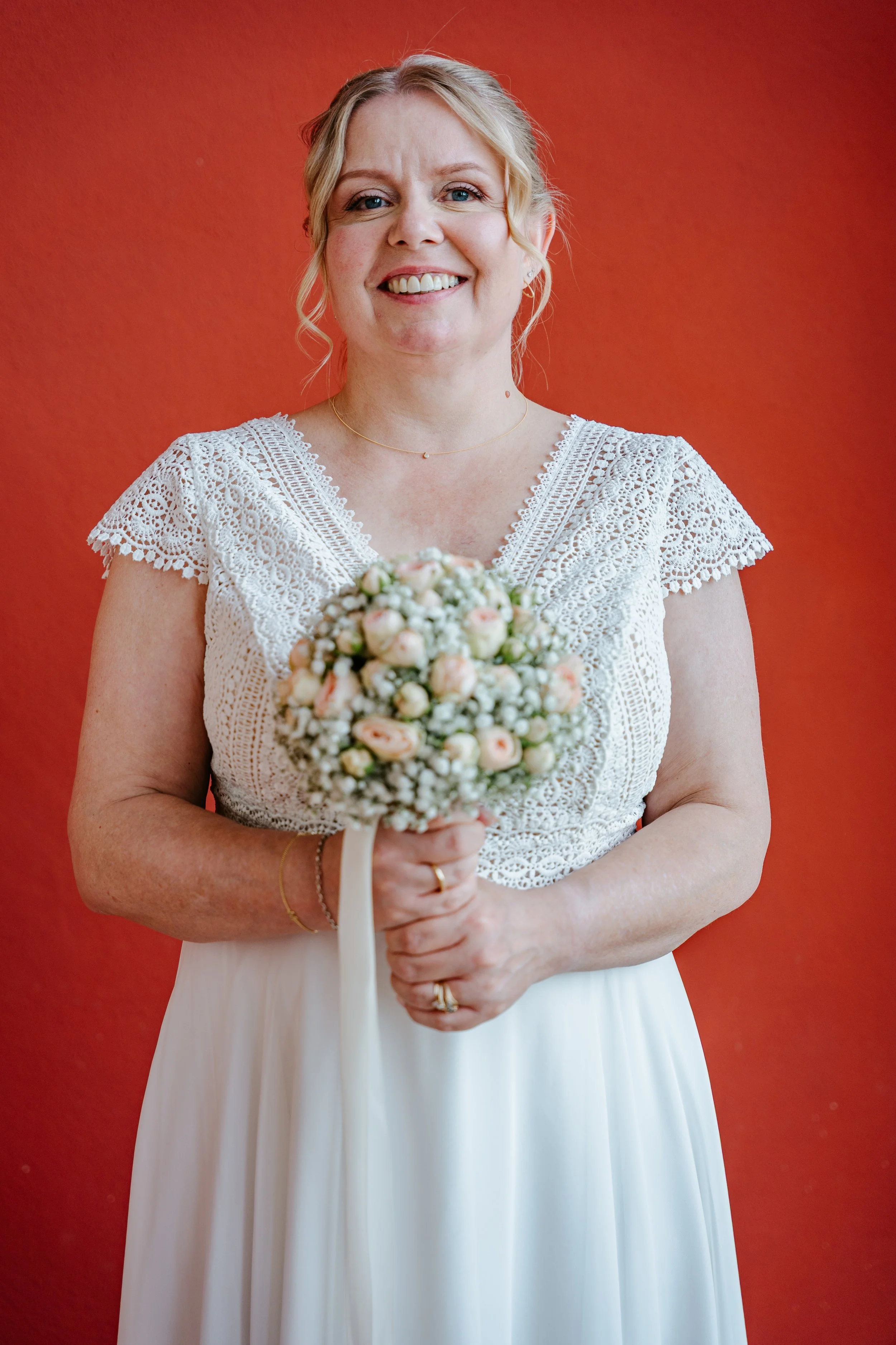 Lächelnde Braut im weißen Spitzenkleid hält einen Blumenstrauß vor rotem Hintergrund.