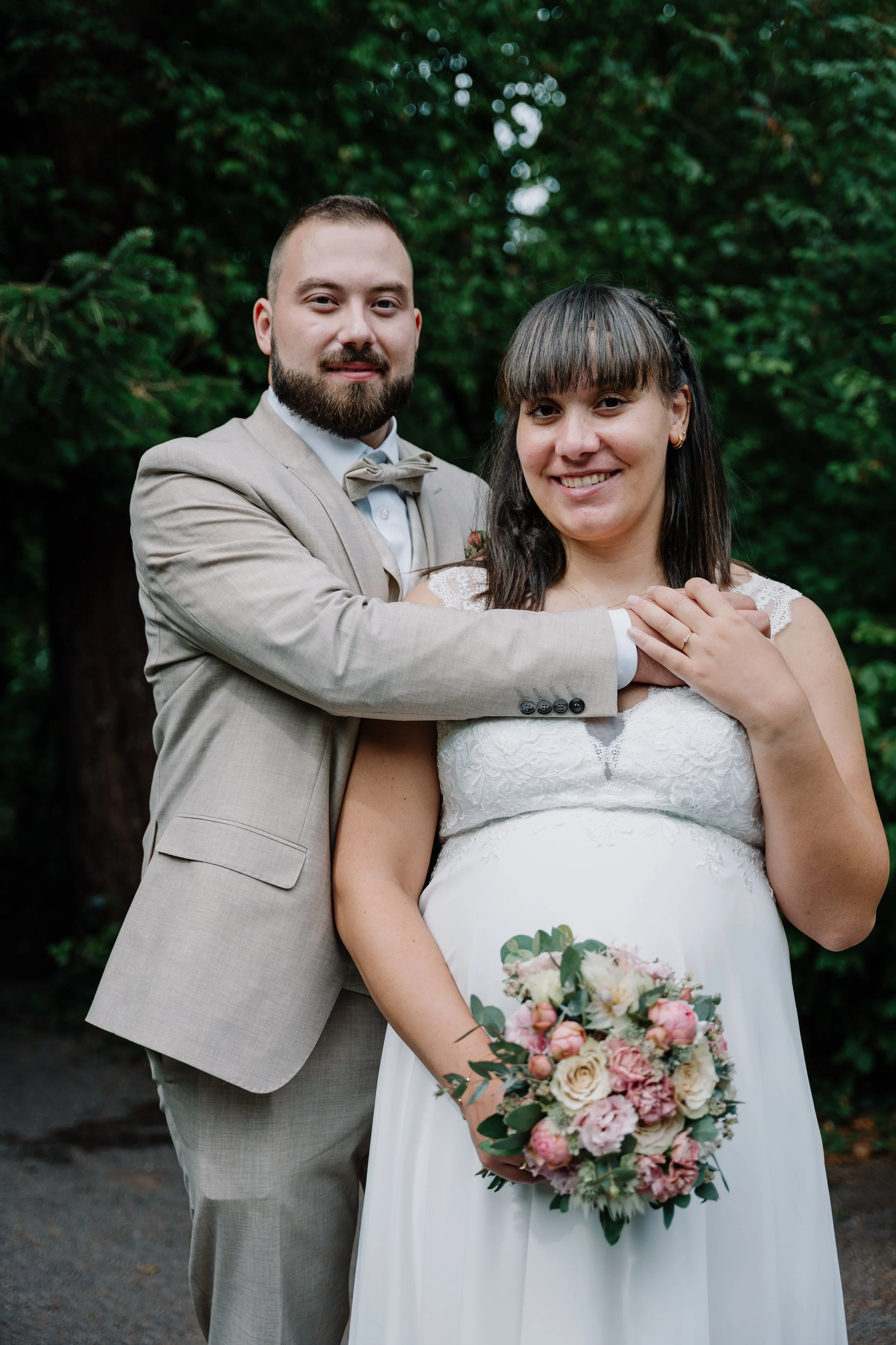 Brautpaar im Wald: lächelnde Braut im weißen Spitzenkleid mit Blumenstrauß und Bräutigam im hellen Anzug
