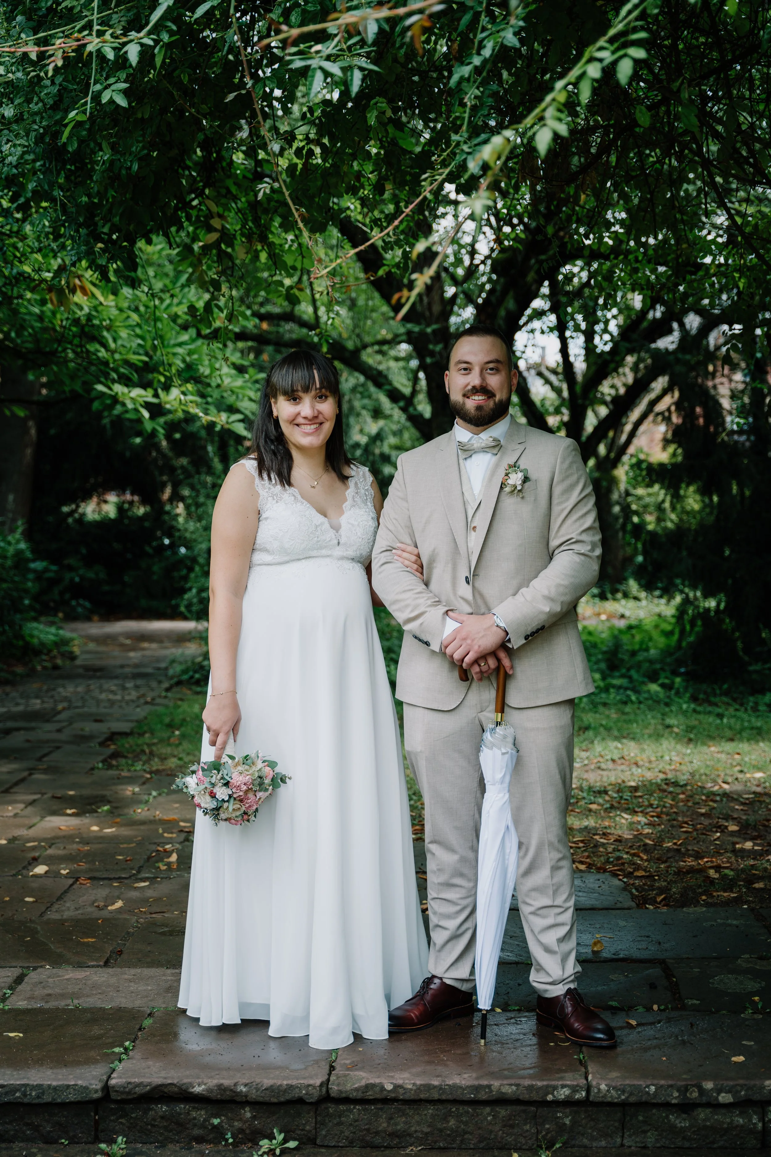 Brautpaar in weißem Hochzeitskleid und hellem Anzug mit Blumenstrauß und Regenschirm in einem grünen Garten